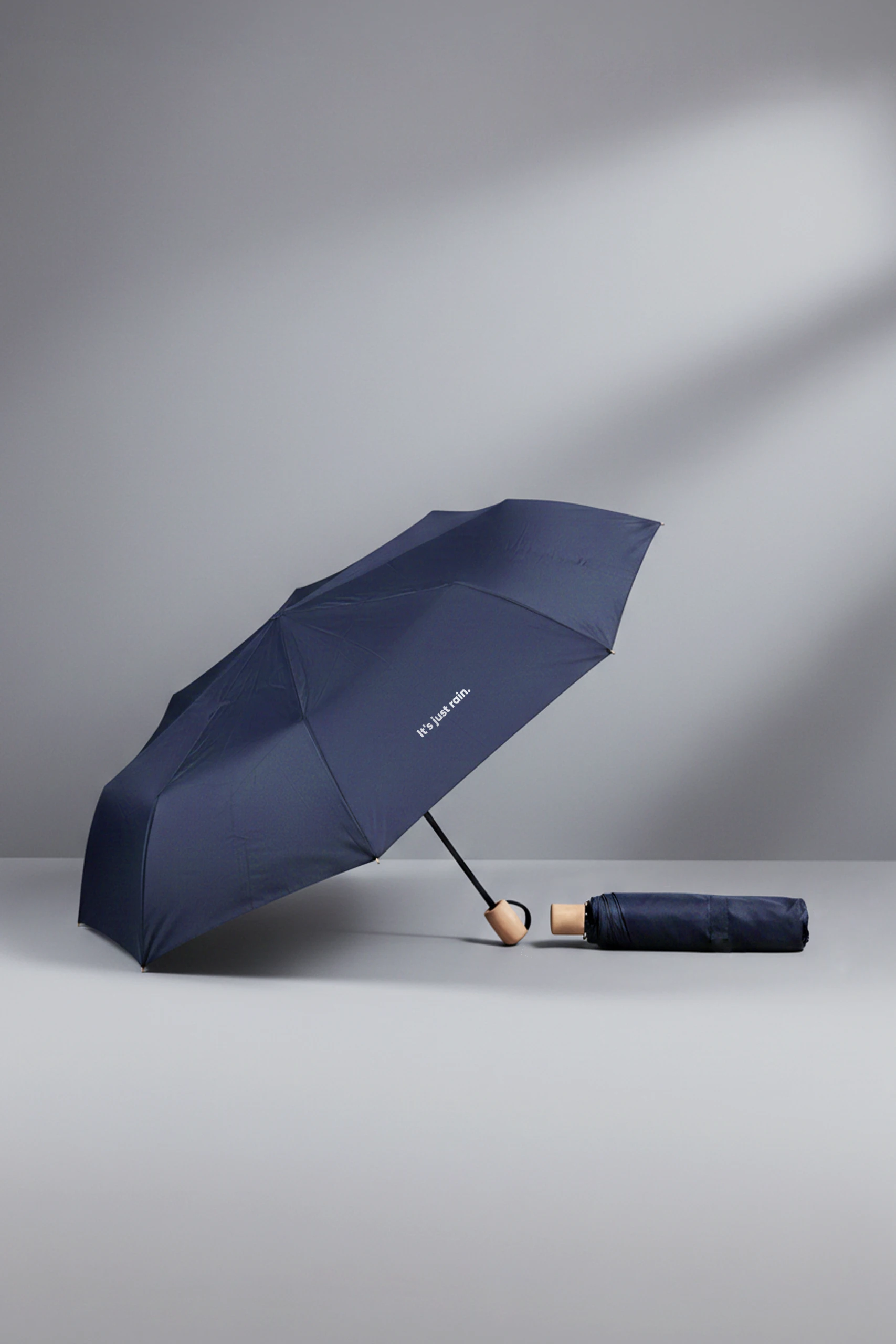 Paraguas abierto personalizable y plegable. Junto con un paraguas cerrado con funda personalizada a todo color. En color azul oscuro. Apoyados en una mesa gris.