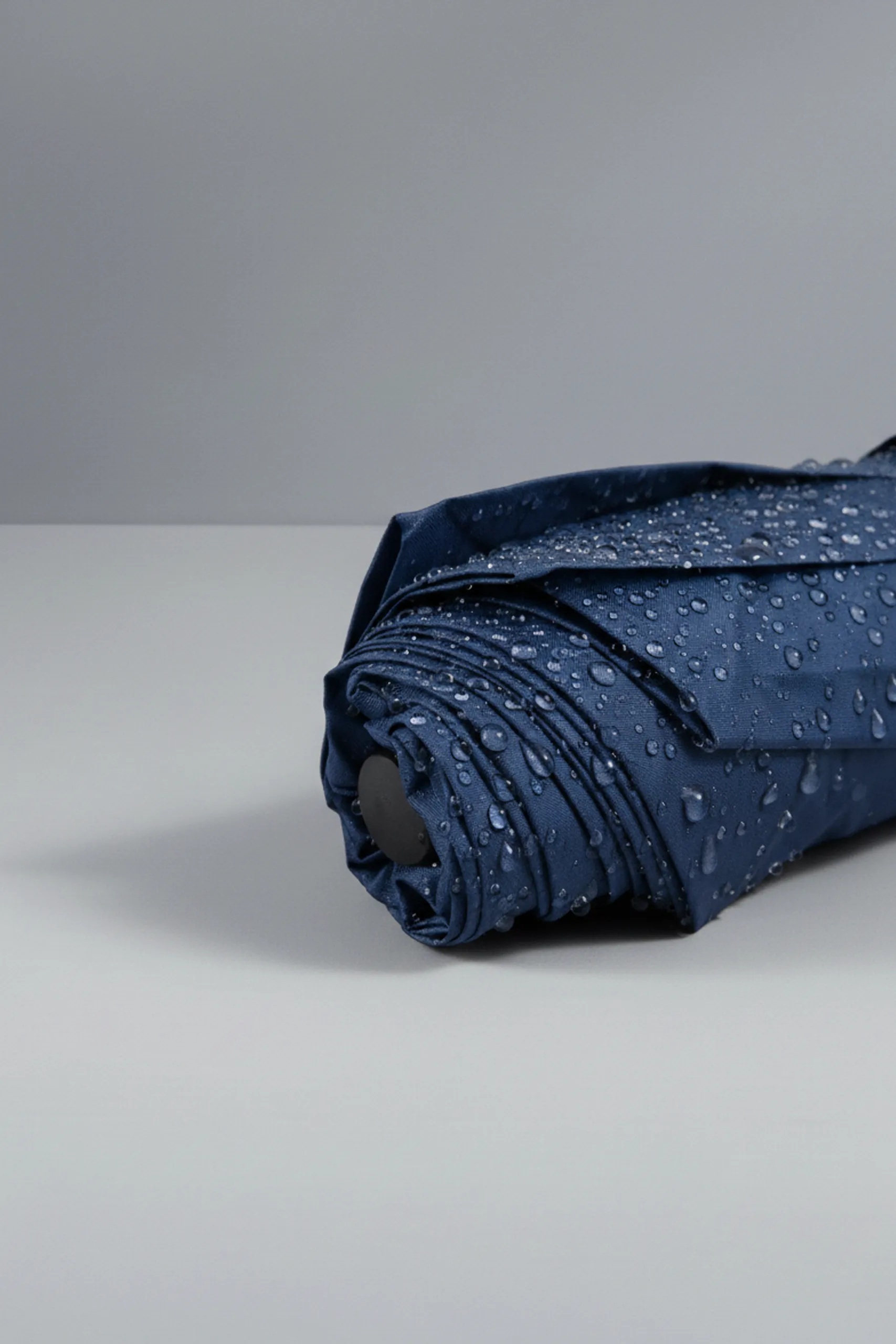 Punta de paraguas plegable personalizado de color azul oscuro , con gotas encima. Apoyado en una mesa gris.