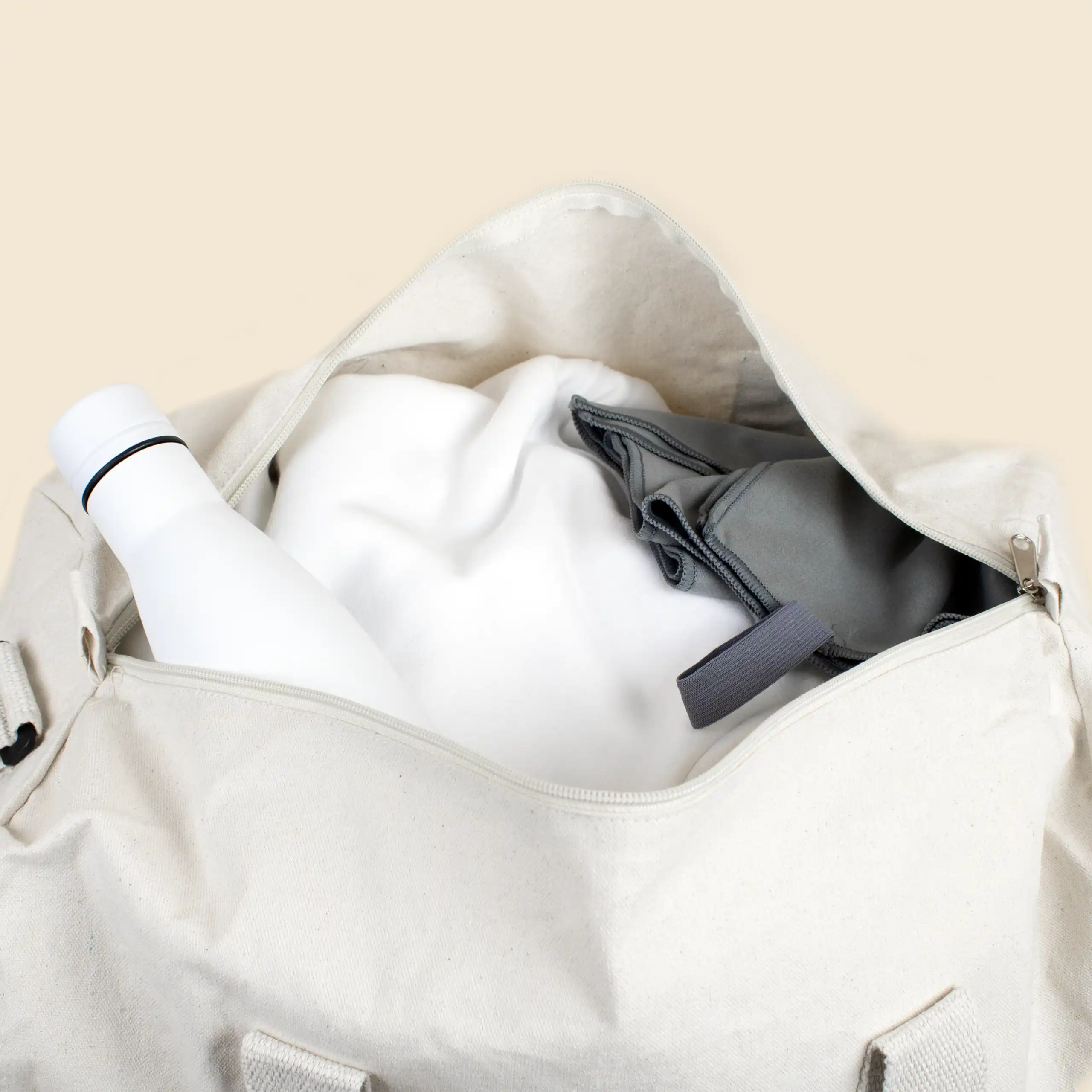 Bolsa de gimnasio personalizada. Abierta con botella blanca, toalla gris de microfibra y textil blanco. De algodón reciclado y color beige. Con personalización a todo color marrón.