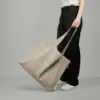 Bolsa tote personalizada GAIA Bolsa tote personalizada. Fabricada en algodón reciclado. De color beige y personalización a todo color. Con una persona sujetándola.