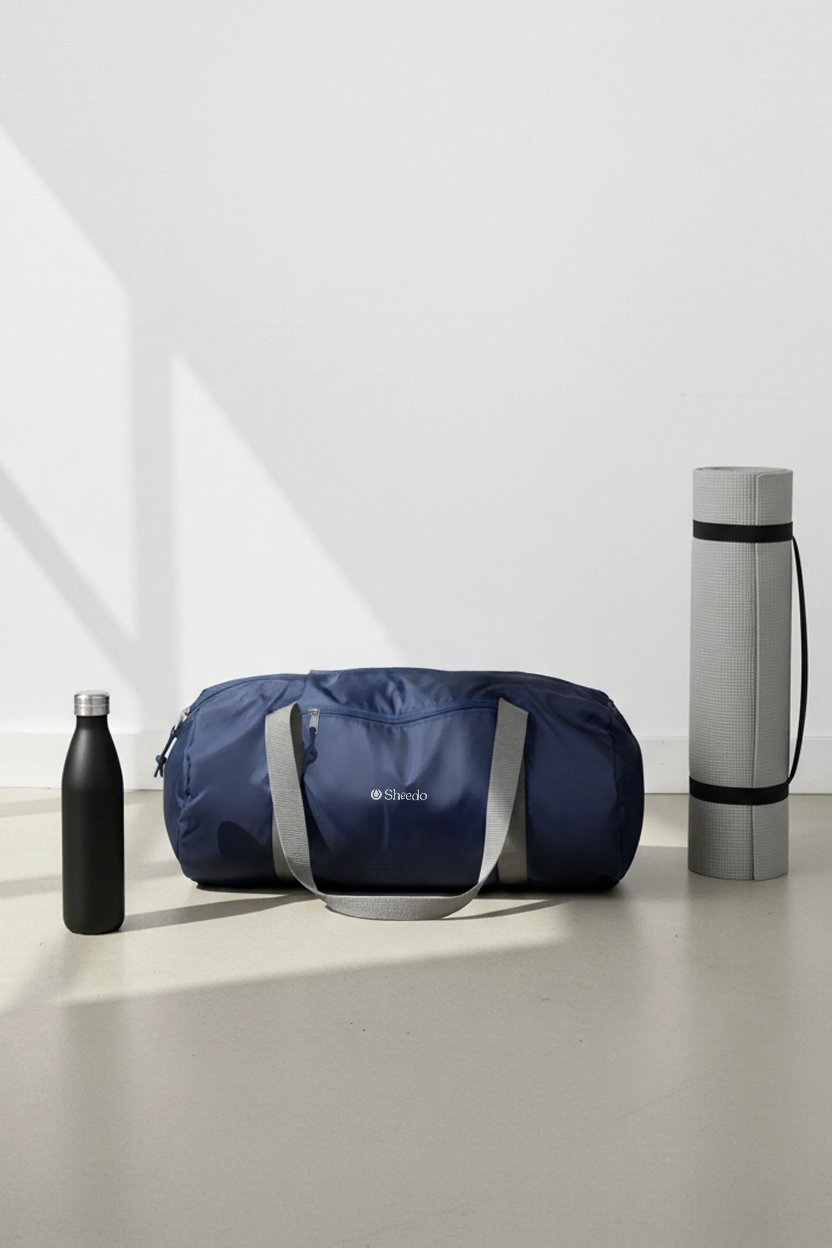 Mochila de deporte personalizada. De color azul marino con asas grises y diseño en blanco. Elaborado a partir de plástico reciclado (RPET). Colocada en el suelo junto con una esterilla gris y una botella reciclada negra mate.