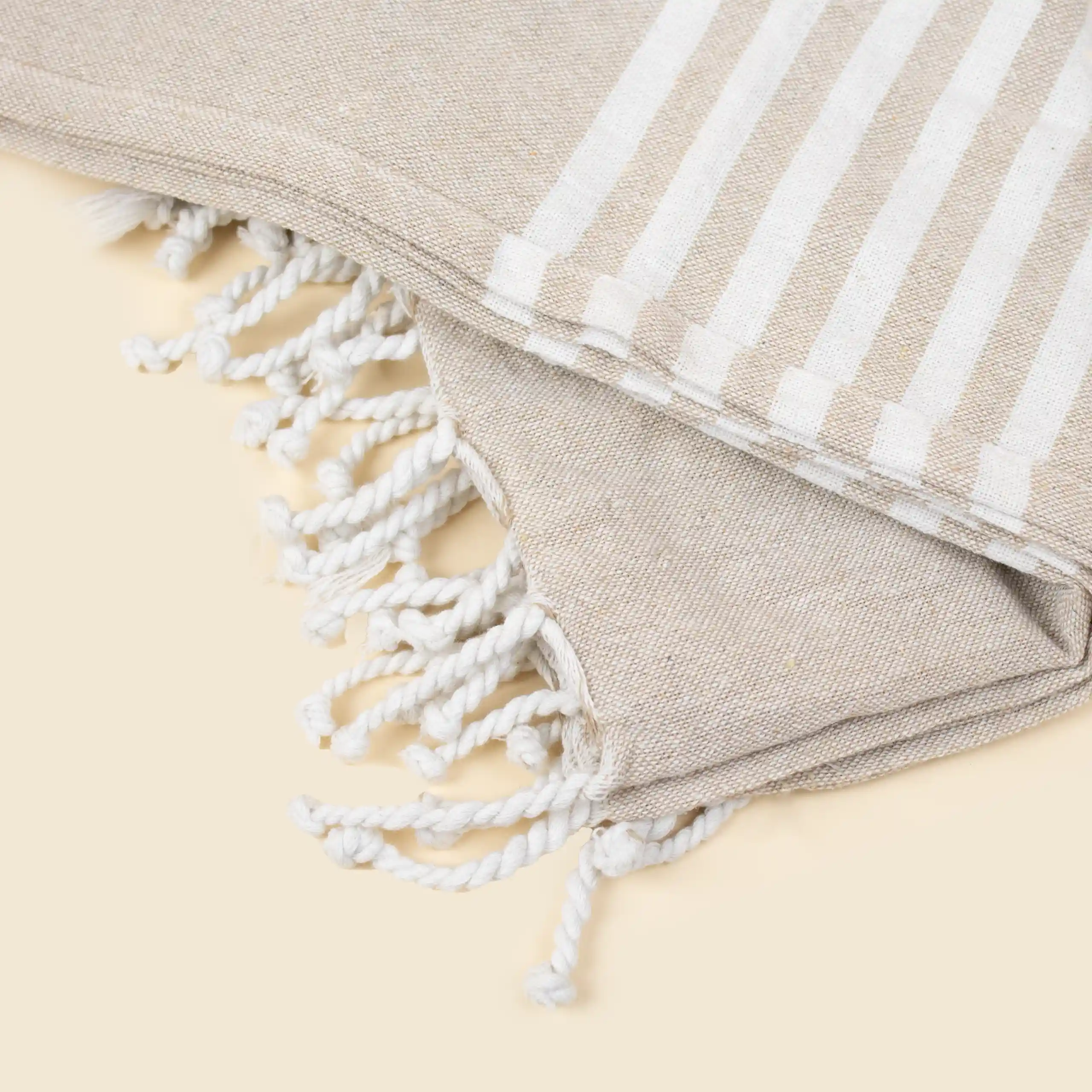 Pareo personalizado, 100% algodón , a rayas de color beige y flecos. Doblado en una mesa beige.
