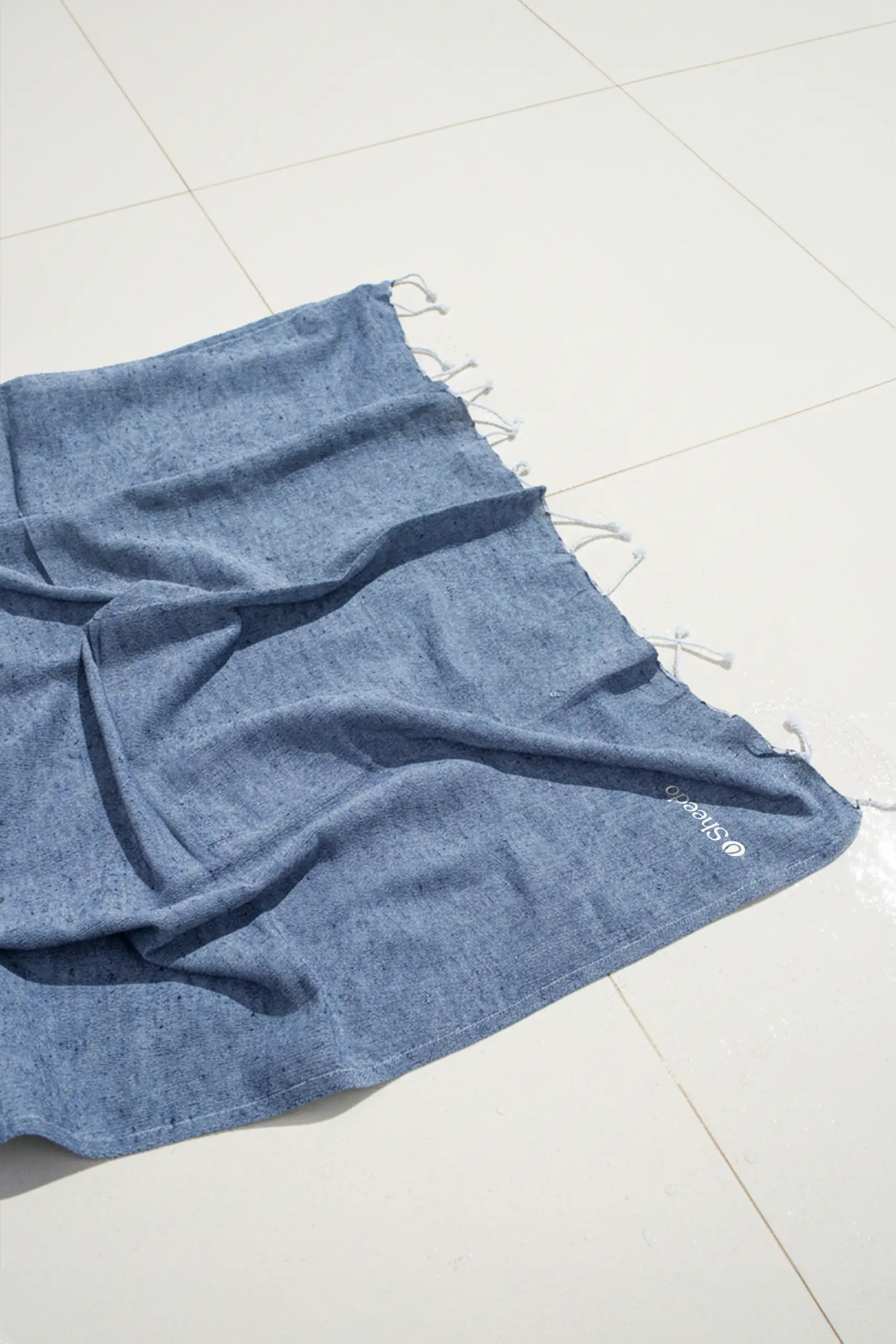 Toalla de playa personalizada con logo blanco. Fabricado de RPET. De color azul y con flecos. Extendida en el suelo.