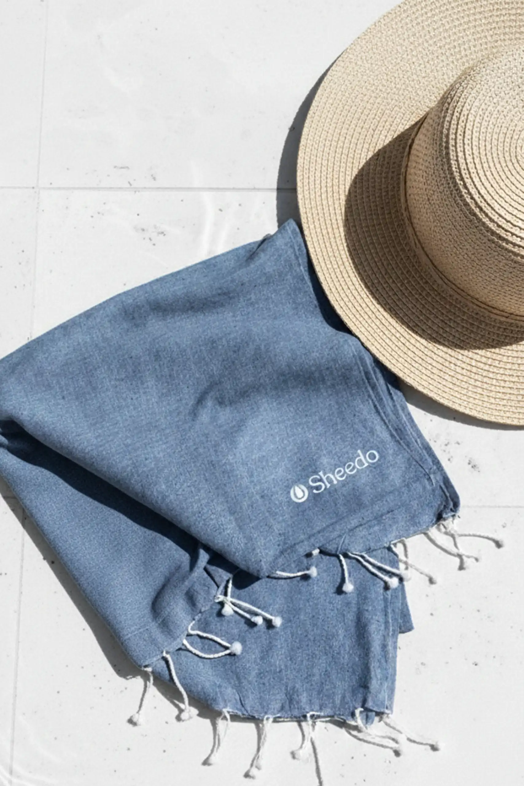 Toalla de playa personalizada con logo blanco. Fabricado de RPET. De color azul y con flecos. Colocada en el suelo con una pamela de mimbre.