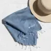 Toalla de playa personalizada_CAPRI Toalla de playa personalizada con logo blanco. Fabricado de RPET. De color azul y con flecos. Colocada en el suelo con una pamela de mimbre.