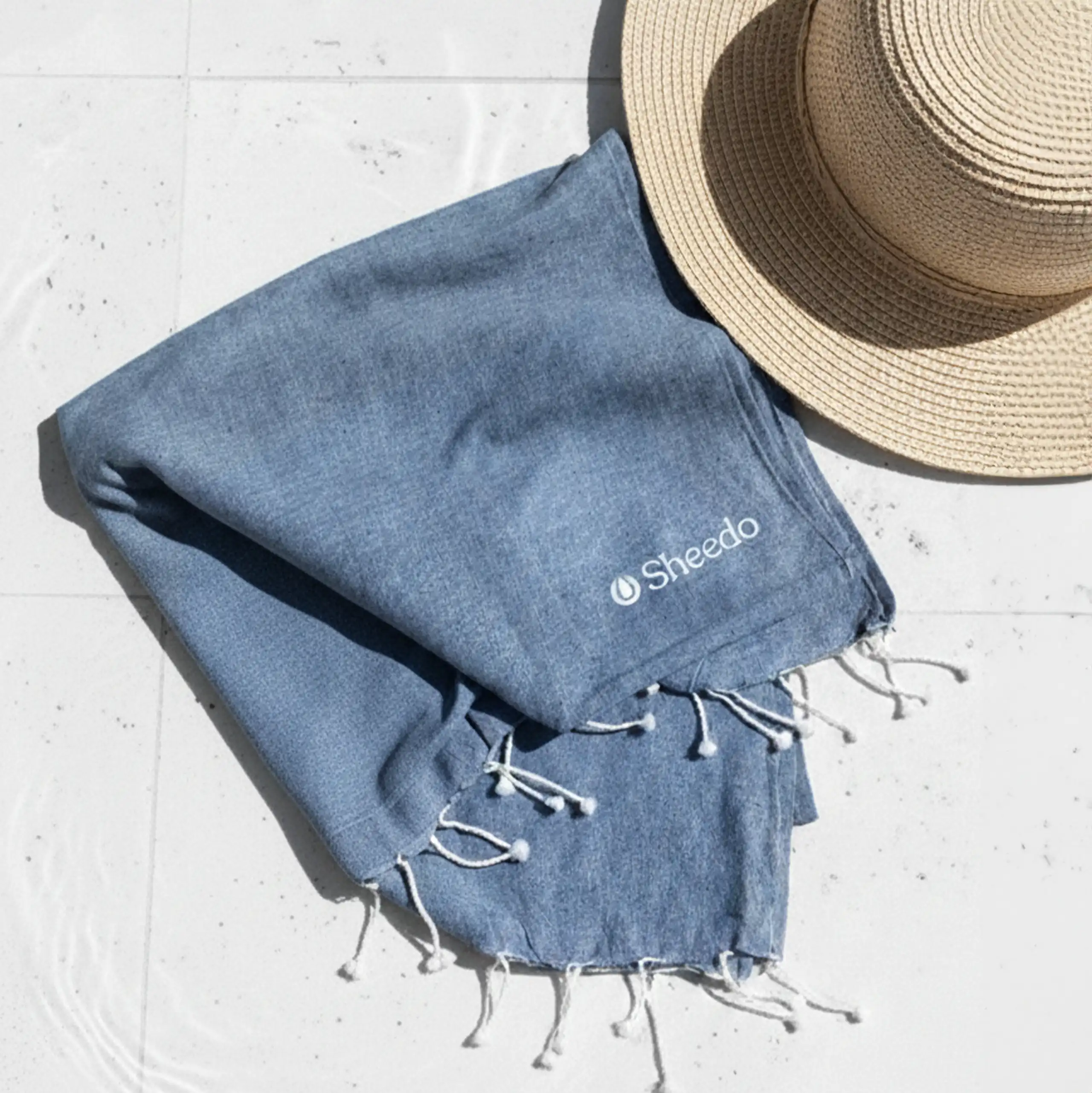 Toalla de playa personalizada_CAPRI Toalla de playa personalizada con logo blanco. Fabricado de RPET. De color azul y con flecos. Colocada en el suelo con una pamela de mimbre.