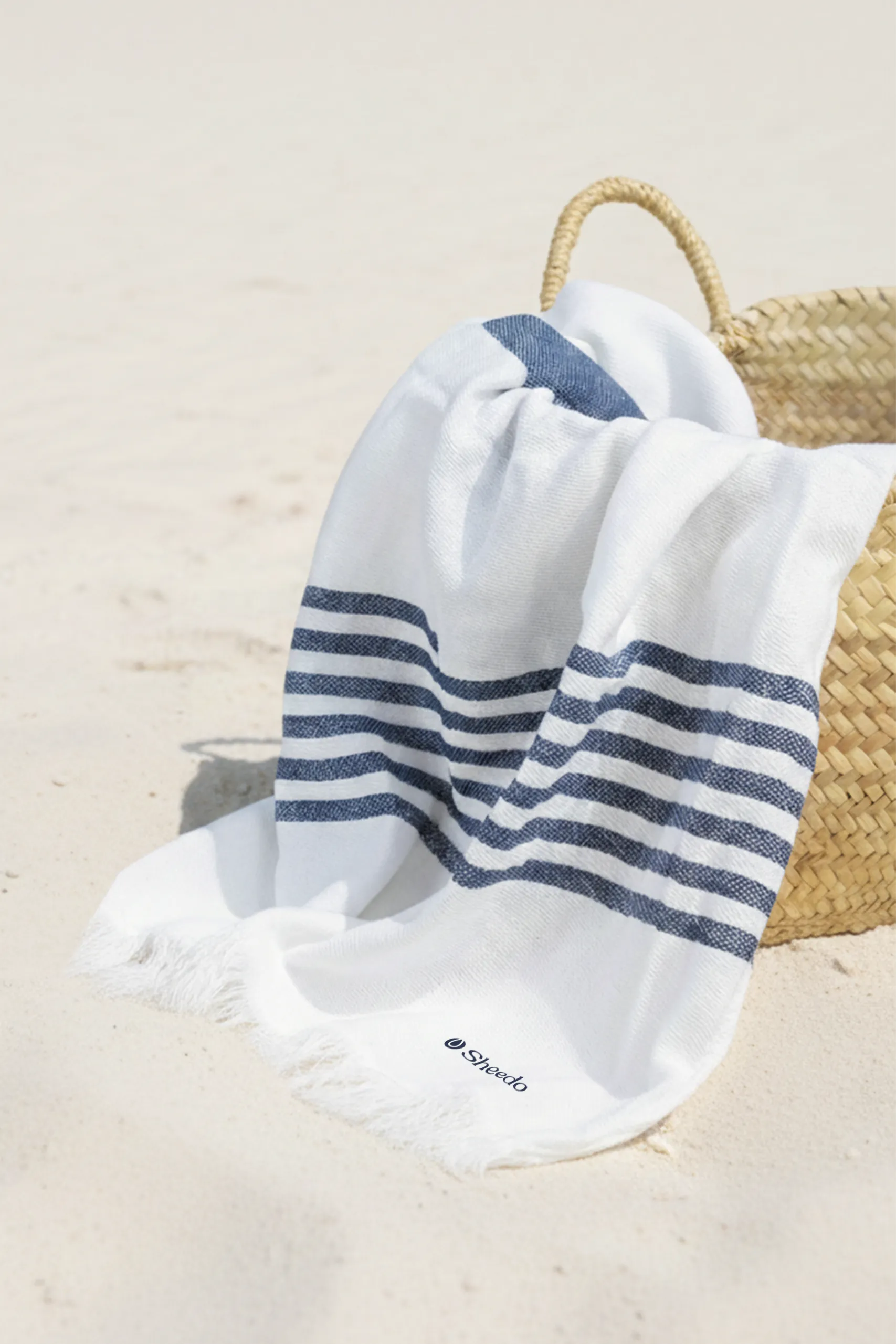 Toalla pareo personalizado. Con flecos y de color azul y blanco a rayas. Colocado en la playa en una cesta de mimbre.