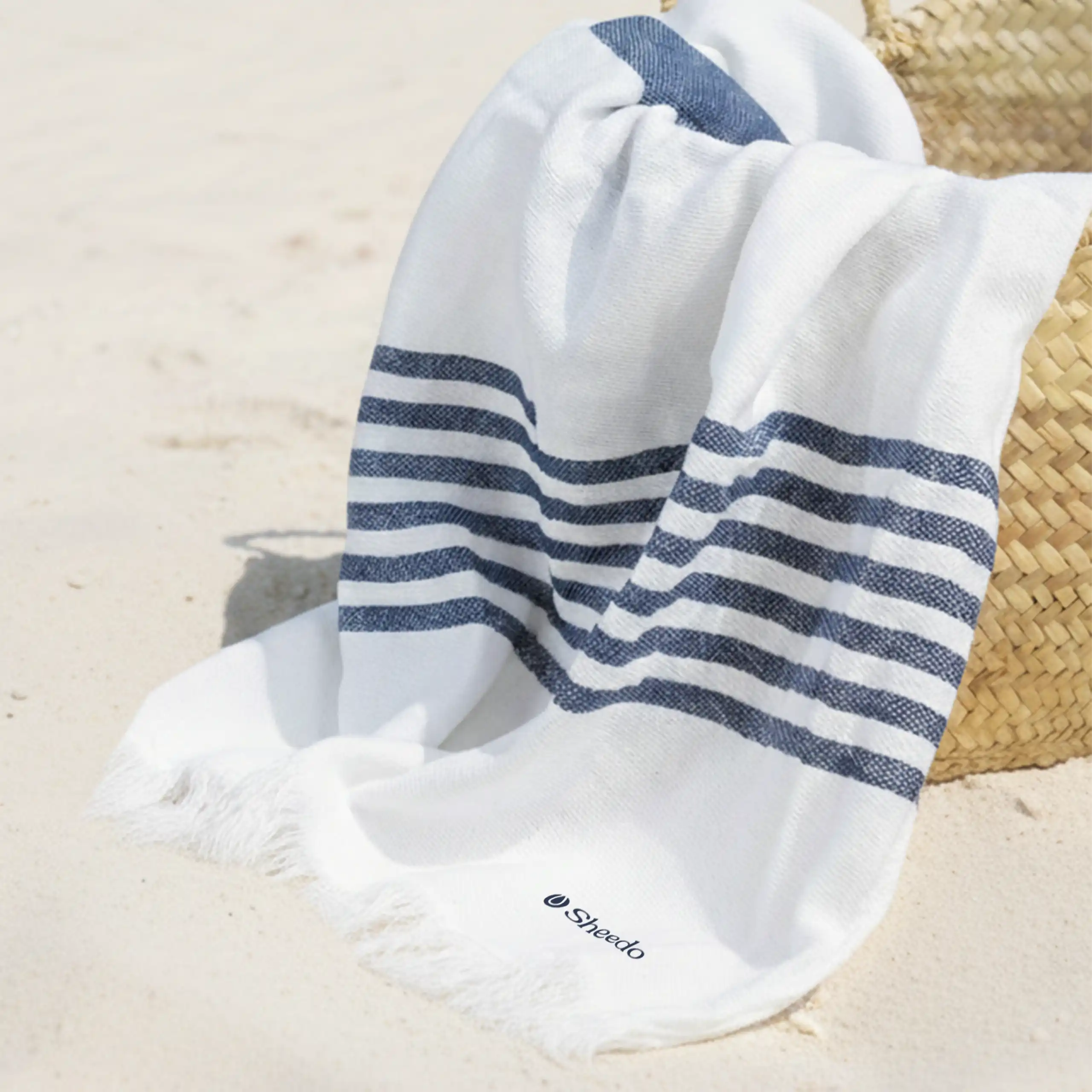 Toalla pareo personalizado. Con flecos y de color azul y blanco a rayas. Colocado en la playa en una cesta de mimbre.