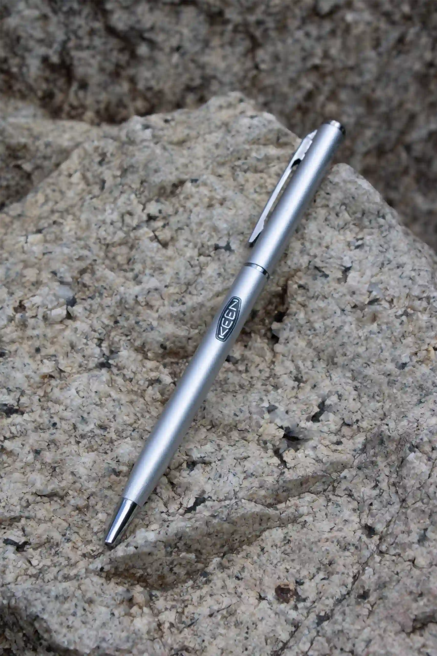 Bolígrafo plateado personalizado con el logotipo de KEEN apoyado sobre una roca