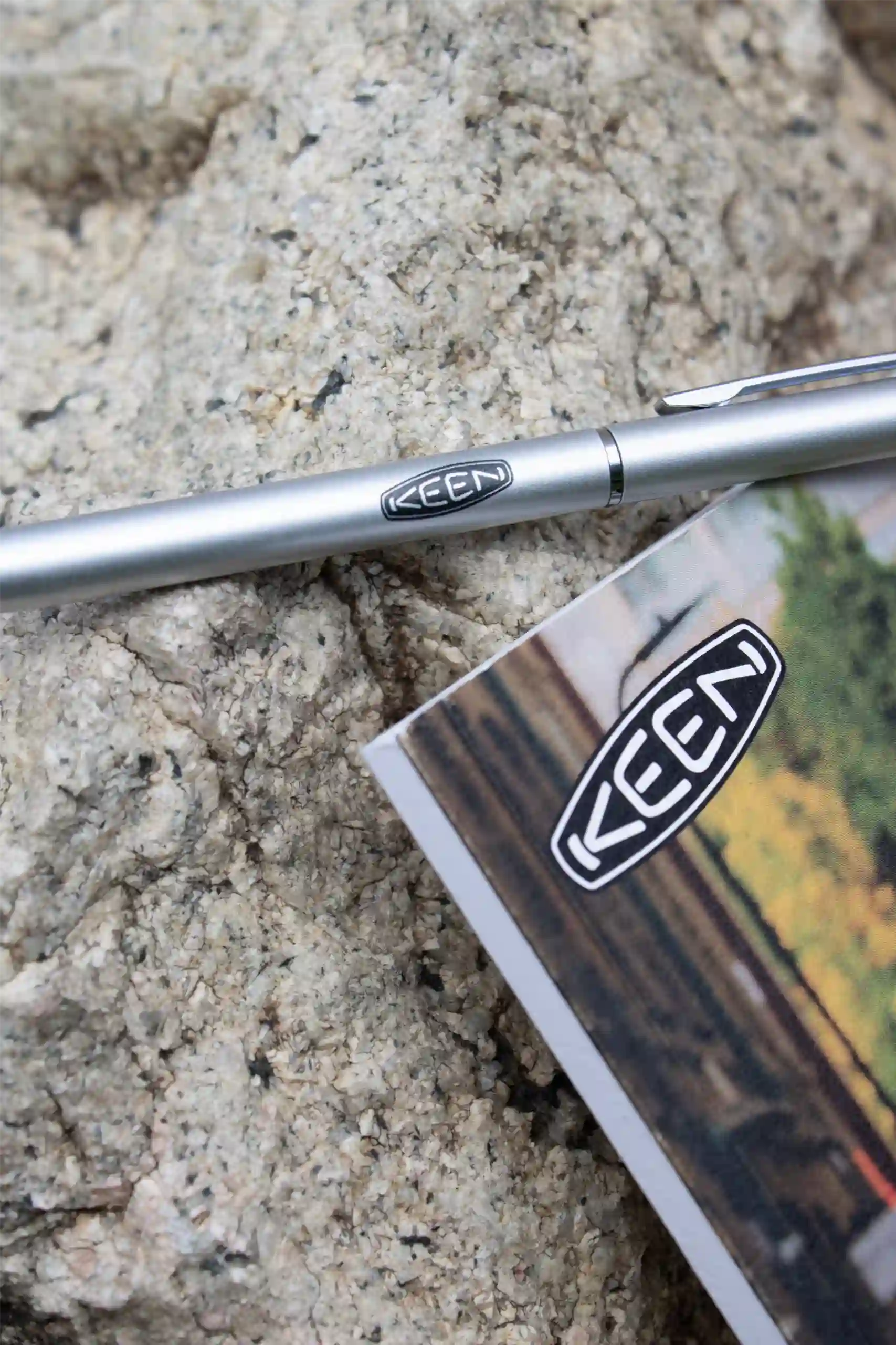 Bolígrafo plateado y cuaderno personalizados con el logotipo de KEEN apoyados sobre una roca
