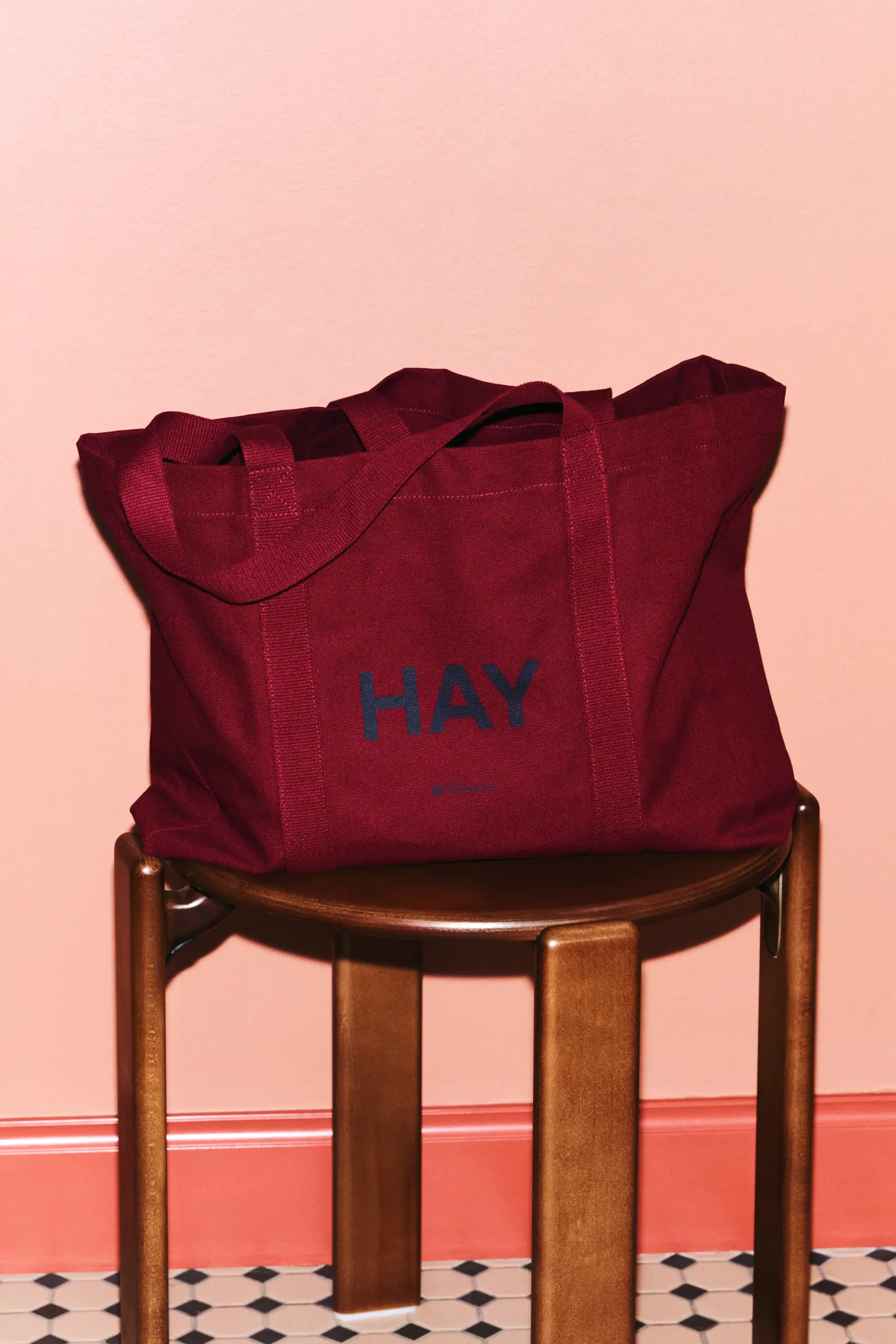 Bolsa de diseño de la marca HAY con impresión a todo color y a una tinta. De color rojo. Apoyada en una silla de madera.