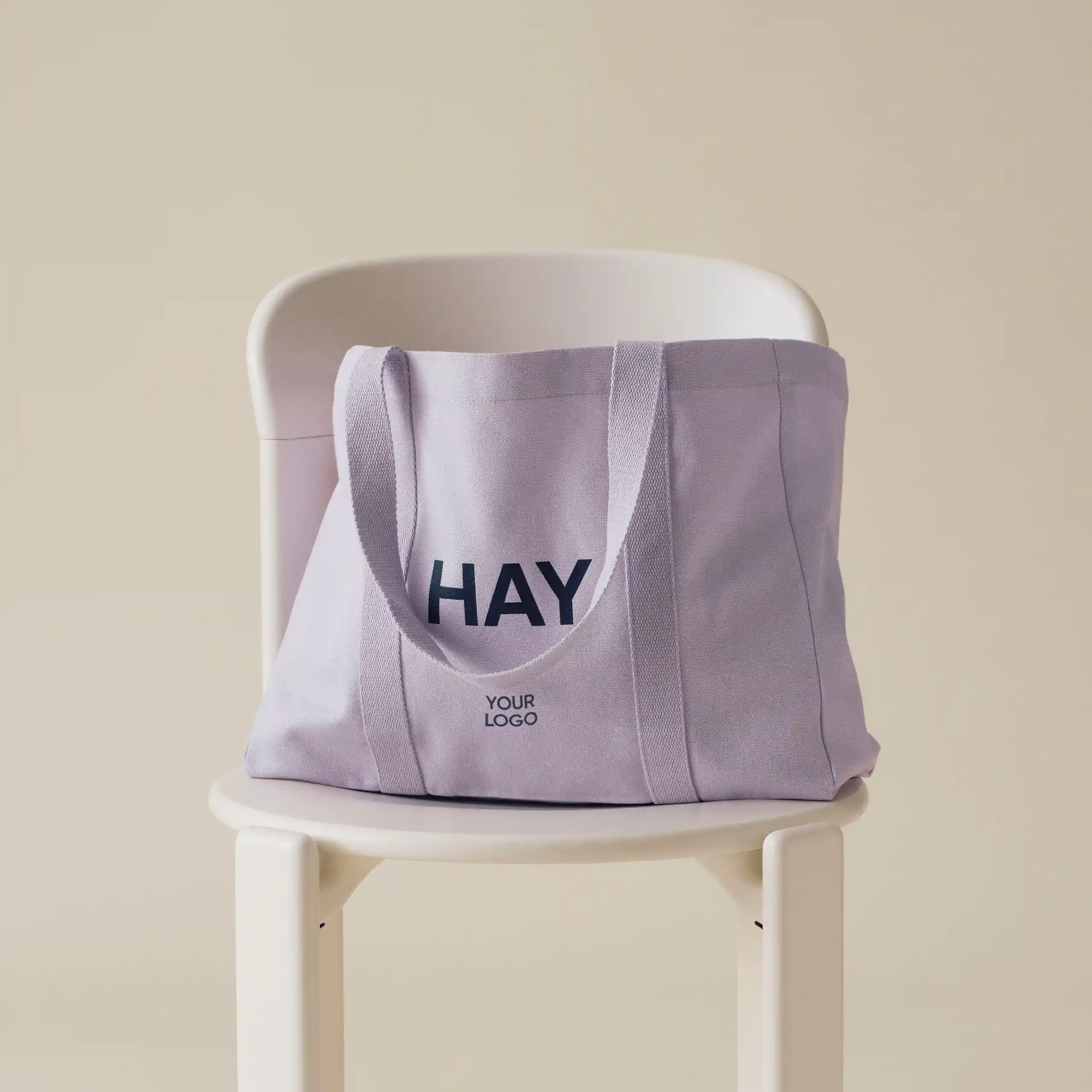 Bolsa de diseño de la marca HAY con impresión a todo color y a una tinta. De color lila. Apoyada en una silla blanca.
