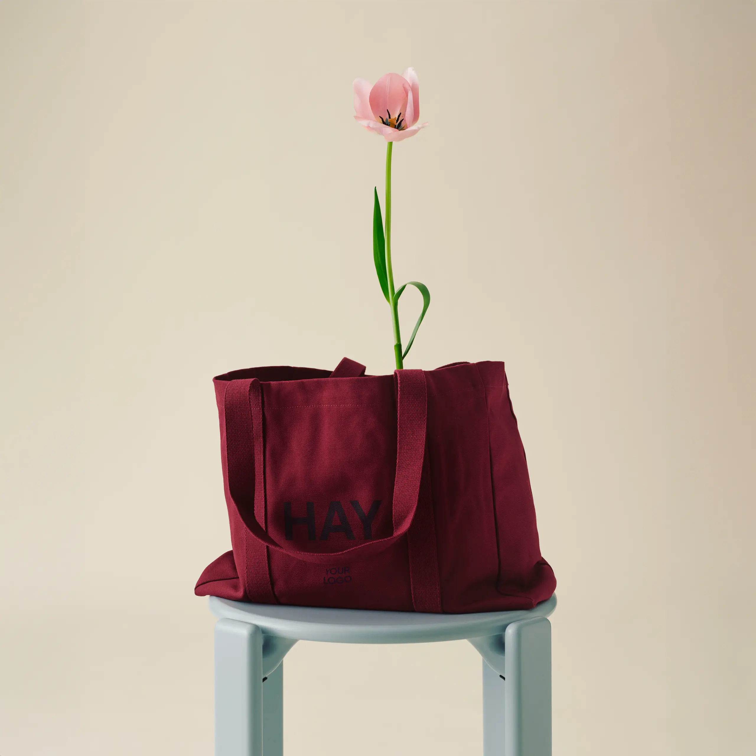 Bolsa de diseño de la marca HAY con impresión a todo color y a una tinta. De color rojo. Apoyada en una silla azul y con una flor.
