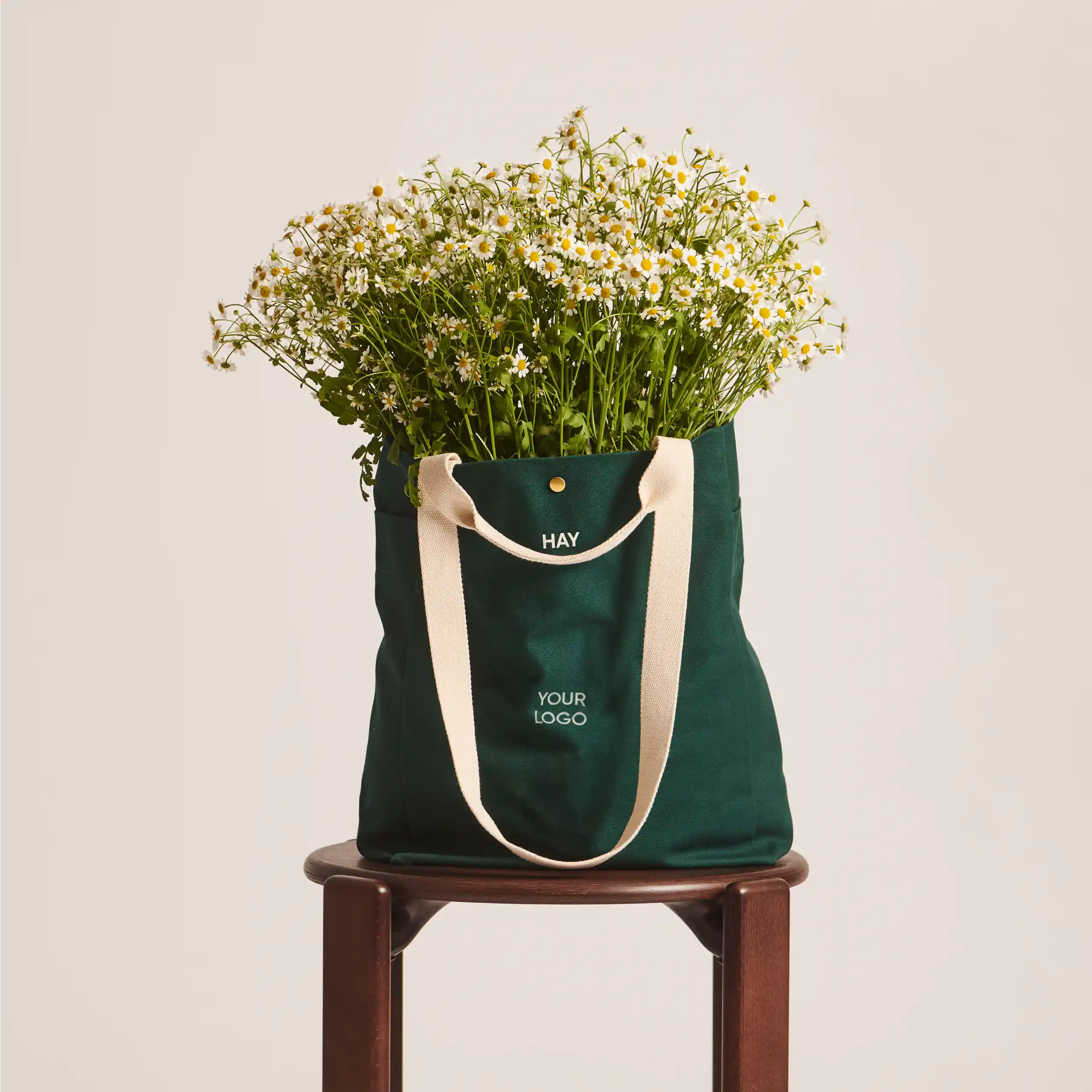 Bolsa tote de algodón orgánico de la marca HAY. Personalizable a todo color y a un color. De color verde y asas beige. Con un ramo de margaritas saliendo de la bolsa.