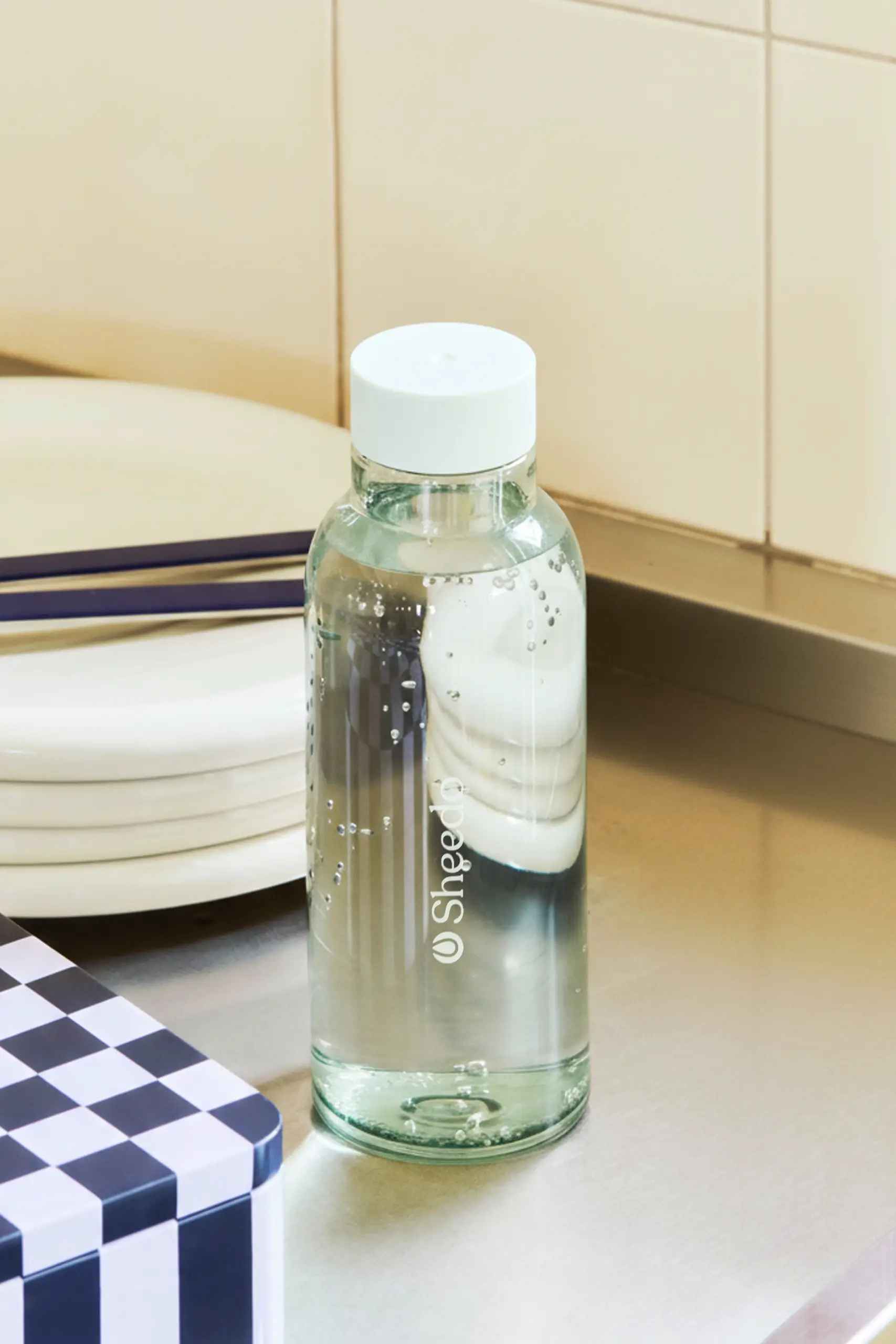 Botella minimalista de la marca HAY. De color azul transparente. Diseñada por el reconocido Naoto Fukasawa . Fabricada en PET reciclado. Colocado en una mesa con cubierto y vajilla.