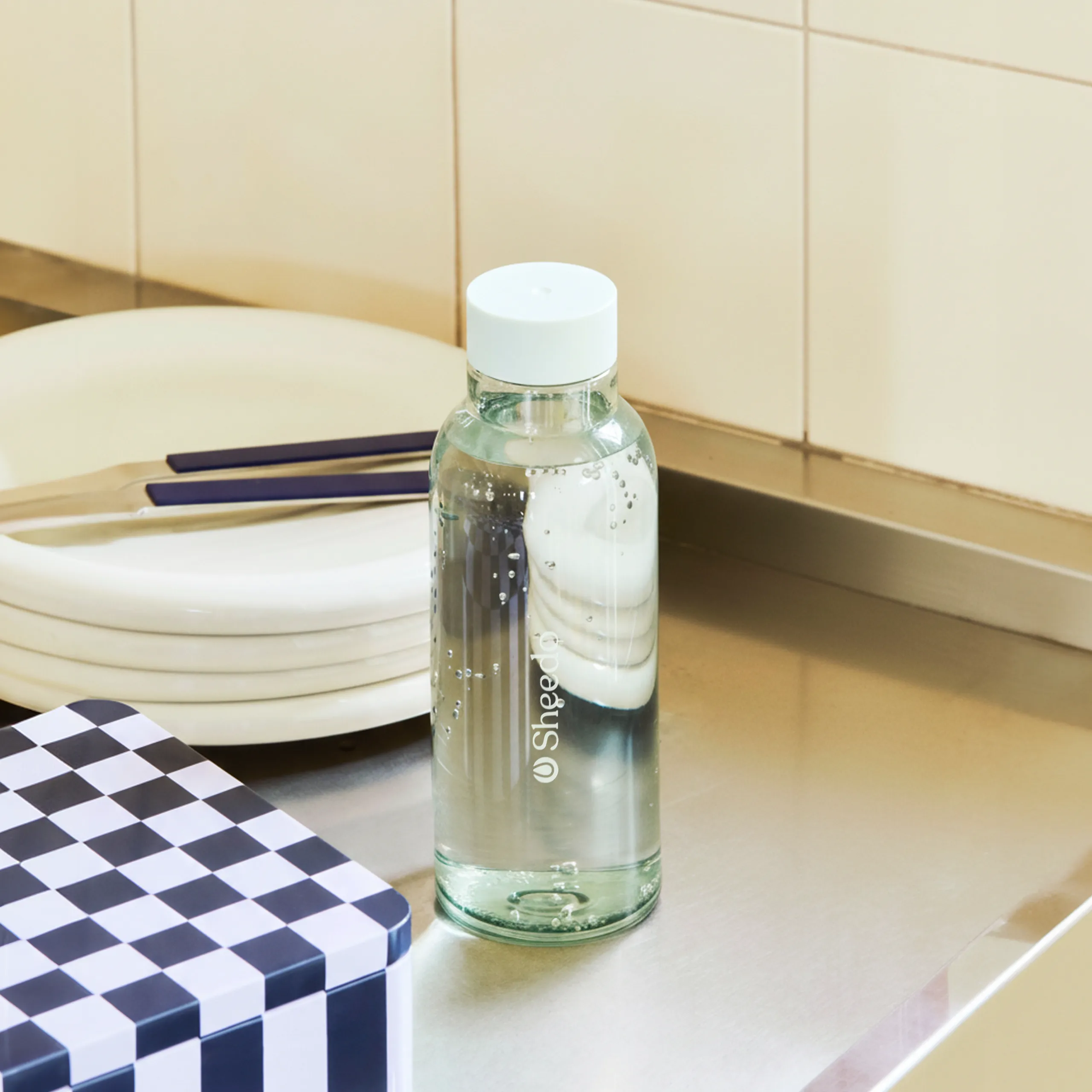 Botella minimalista de la marca HAY. De color azul transparente. Diseñada por el reconocido Naoto Fukasawa . Fabricada en PET reciclado. Colocado en una mesa con cubierto y vajilla.