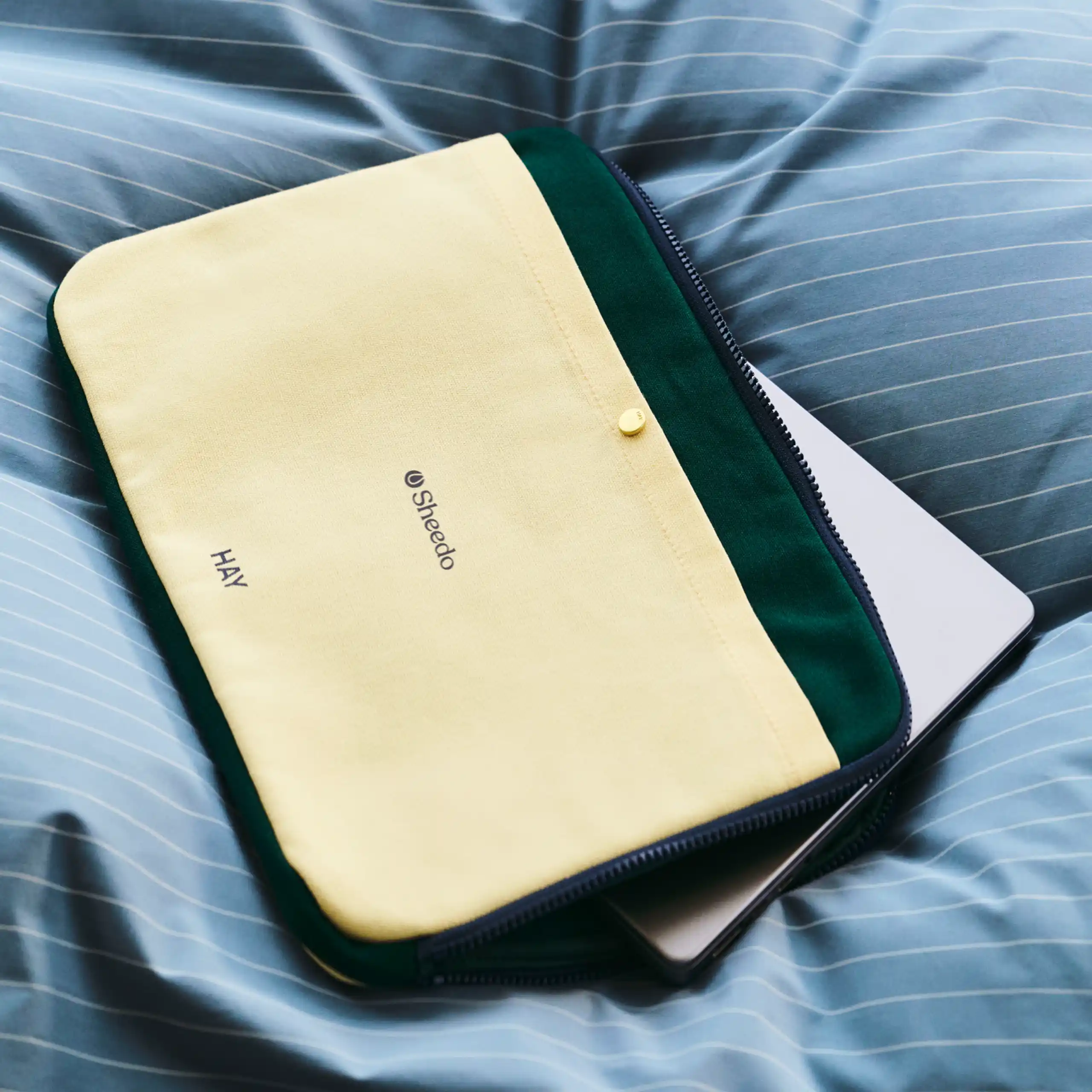 Funda de portátil de la marca HAY. Con botón dorado y cremallera. Combinado de colores amarillo y verde. En una cama con edredón azul.