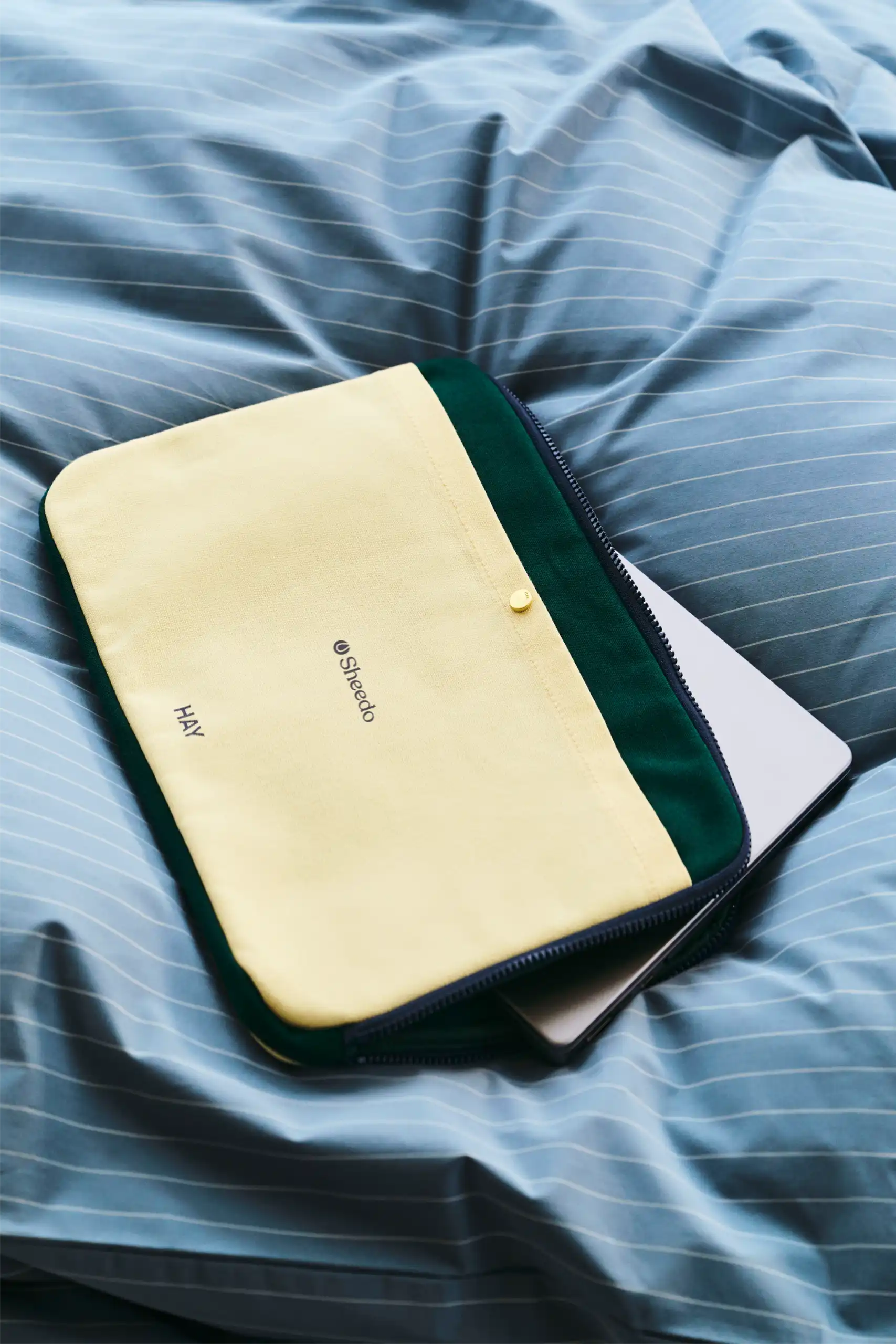 Funda de portátil de la marca HAY. Con botón dorado y cremallera. Combinado de colores amarillo y verde. En una cama con edredón azul.