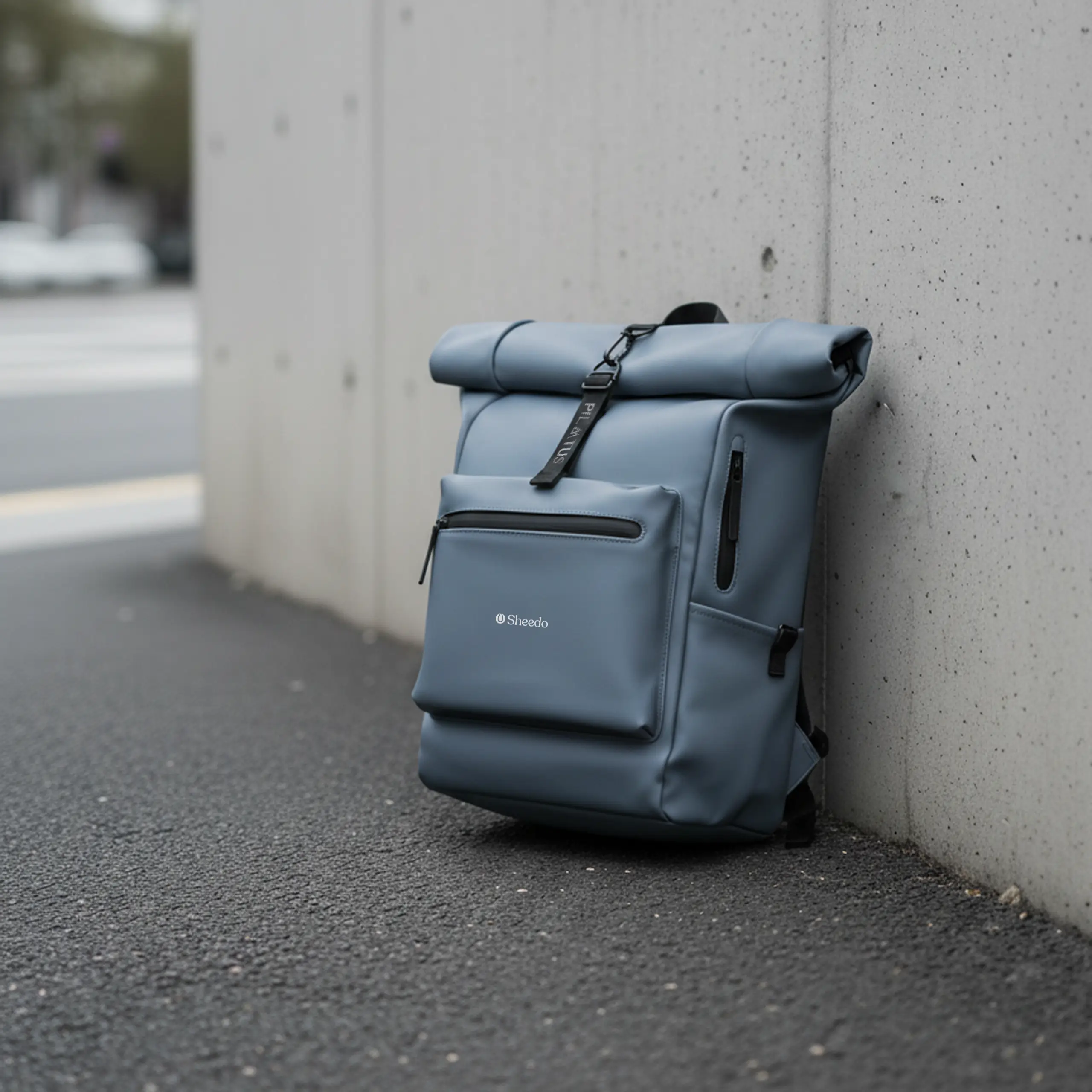 Mochila urbana - Pilatus Urban Mochila urbana y enrollable de la marca Pilatus , de color azul y con personalización a 1 tinta. Colocada en una acera y apoyada en una pared de cemento.