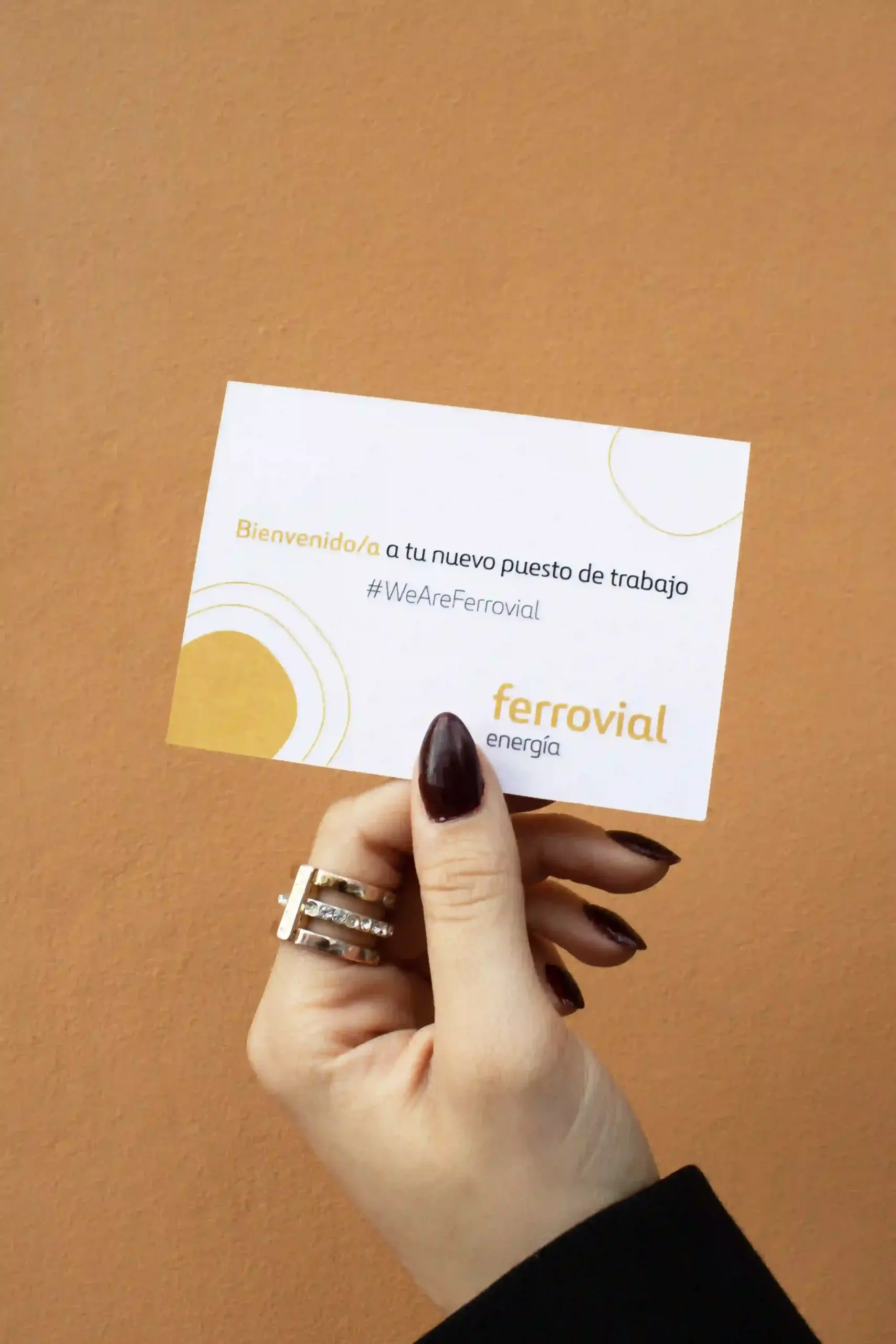 Mano mostrando una tarjeta de visita de la marca Ferrovial sobre un fondo de pared naranja