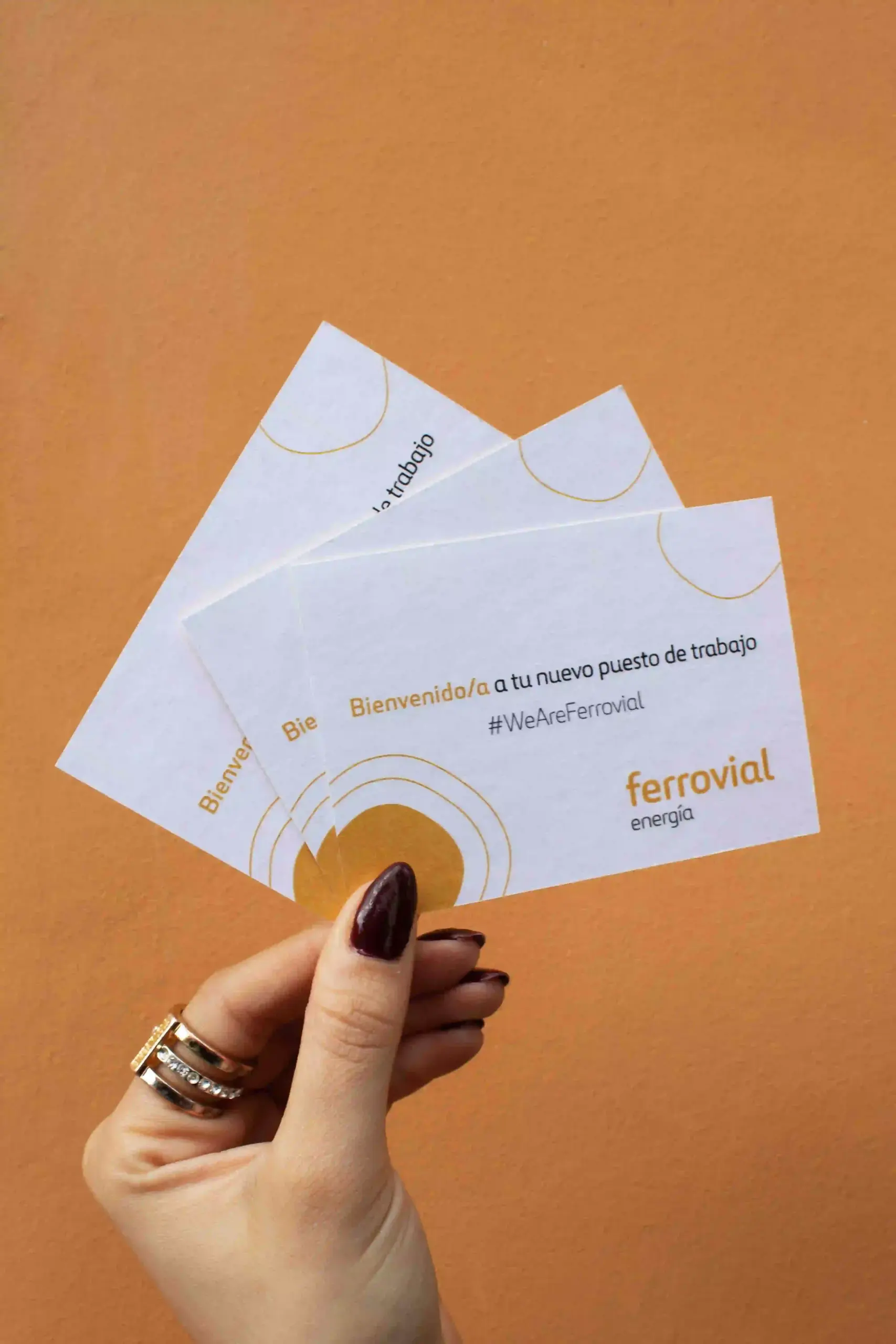Mano mostrando 3 tarjetas de visita de la marca Ferrovial sobre un fondo de pared naranja
