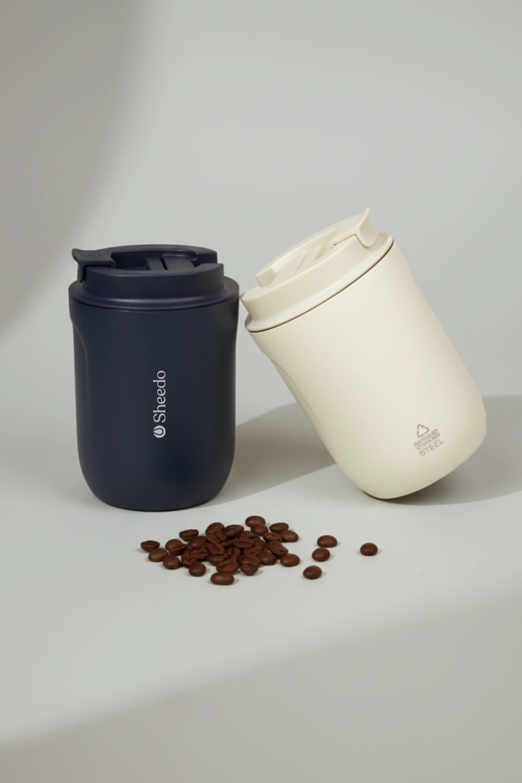 Un vaso térmico azul y otro beige personalizados con el logo de 'Sheedo', junto a un puñado de granos de café