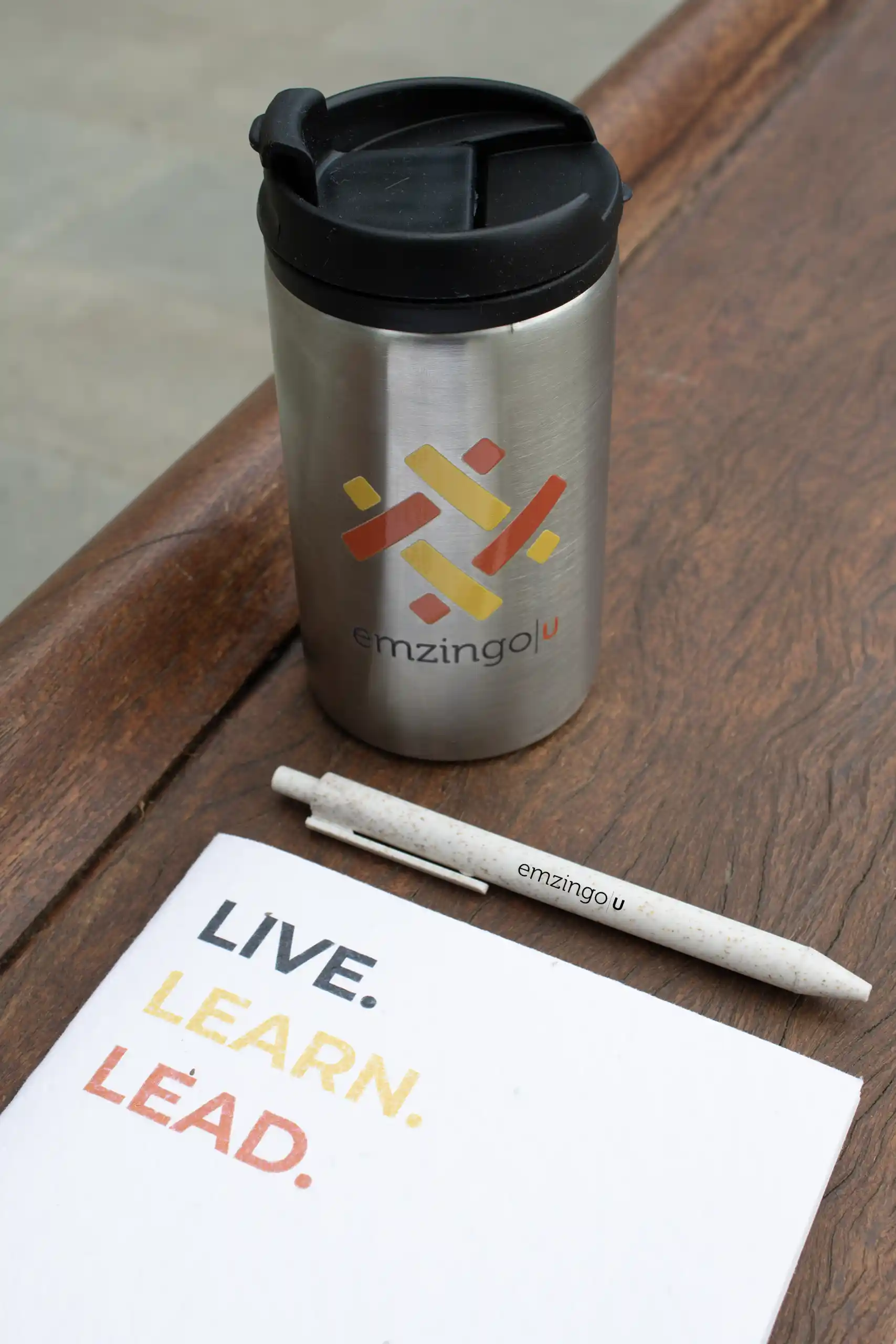 Taza térmica plateada con tapa negra, junto a un cuaderno blanco y un bolígrafo beige, personalizados para la marca Emzingo,