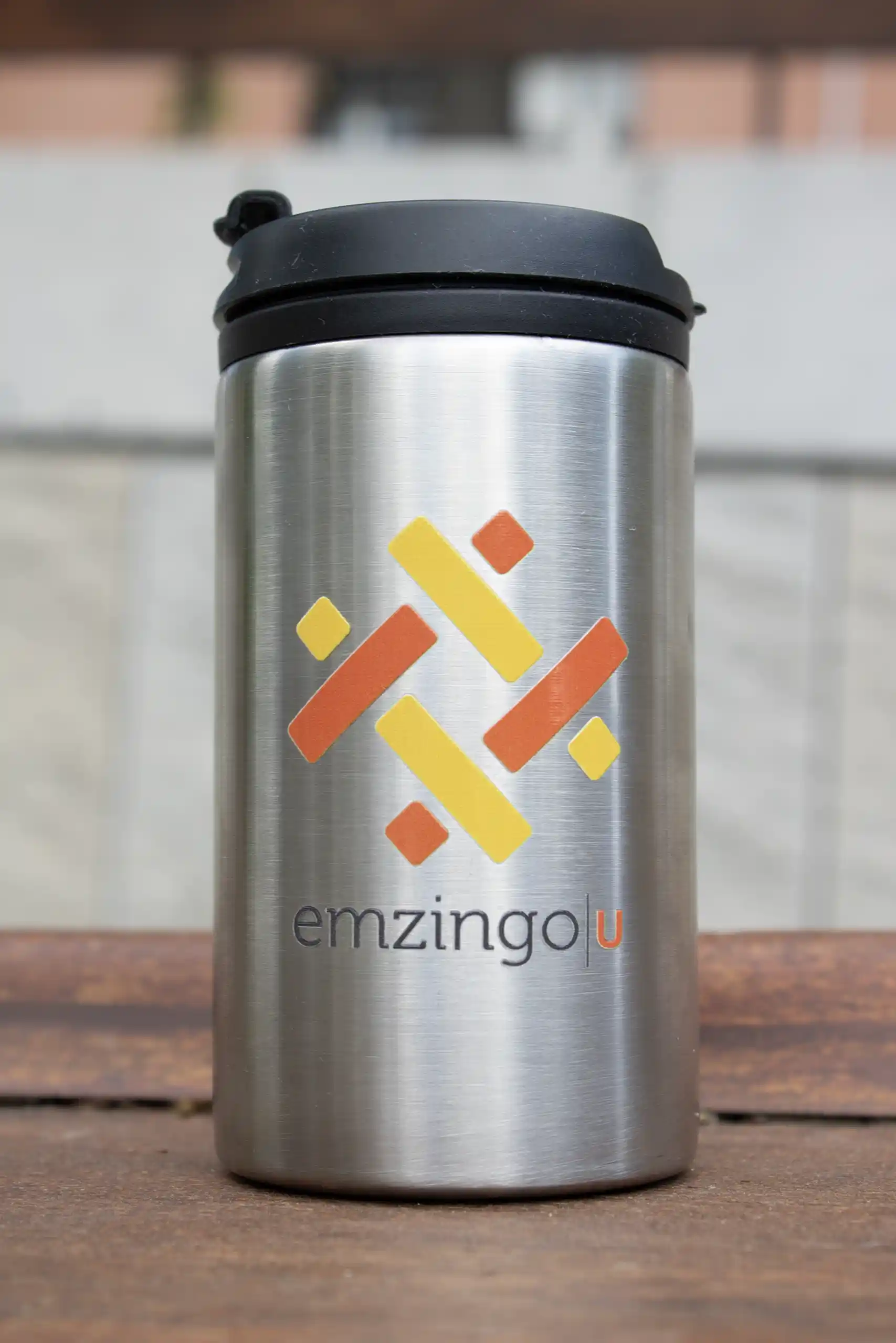 Taza térmica plateada personalizada con el logotipo de la marca Emzingo, apoyada sobre una superficie de madera