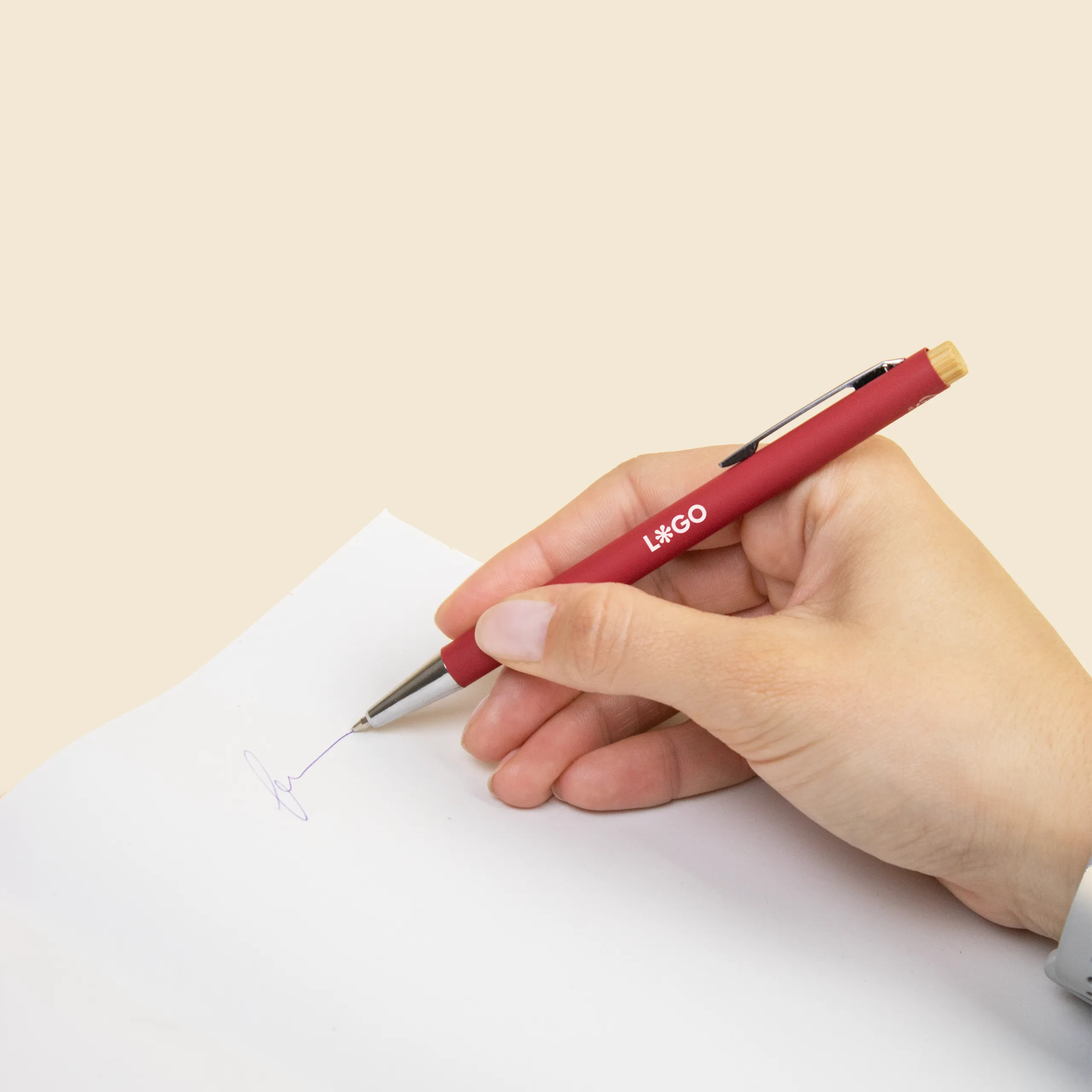 Mano escribiendo en un papel con un bolígrafo rojo de aluminio reciclado y pulsador de bambú, personalizado con la palabra LOGO en blanco