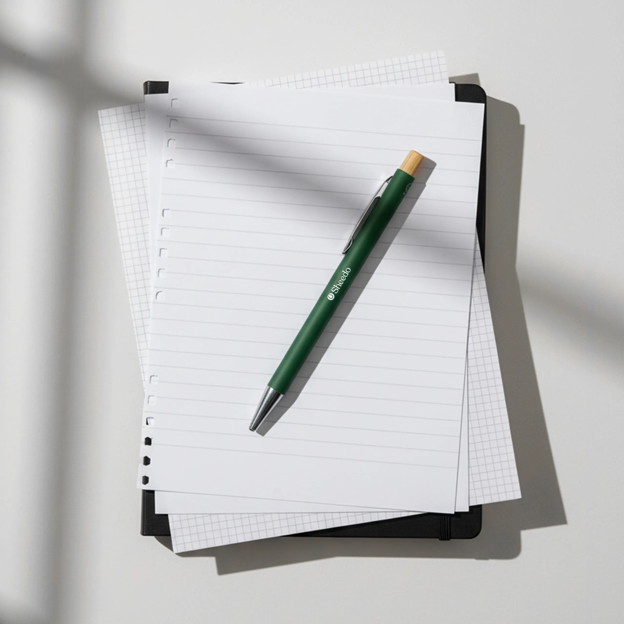 Bolígrafo verde de aluminio reciclado y pulsador de bambú, personalizado con el logo de Sheedo, colocado sobre unos folios blancos