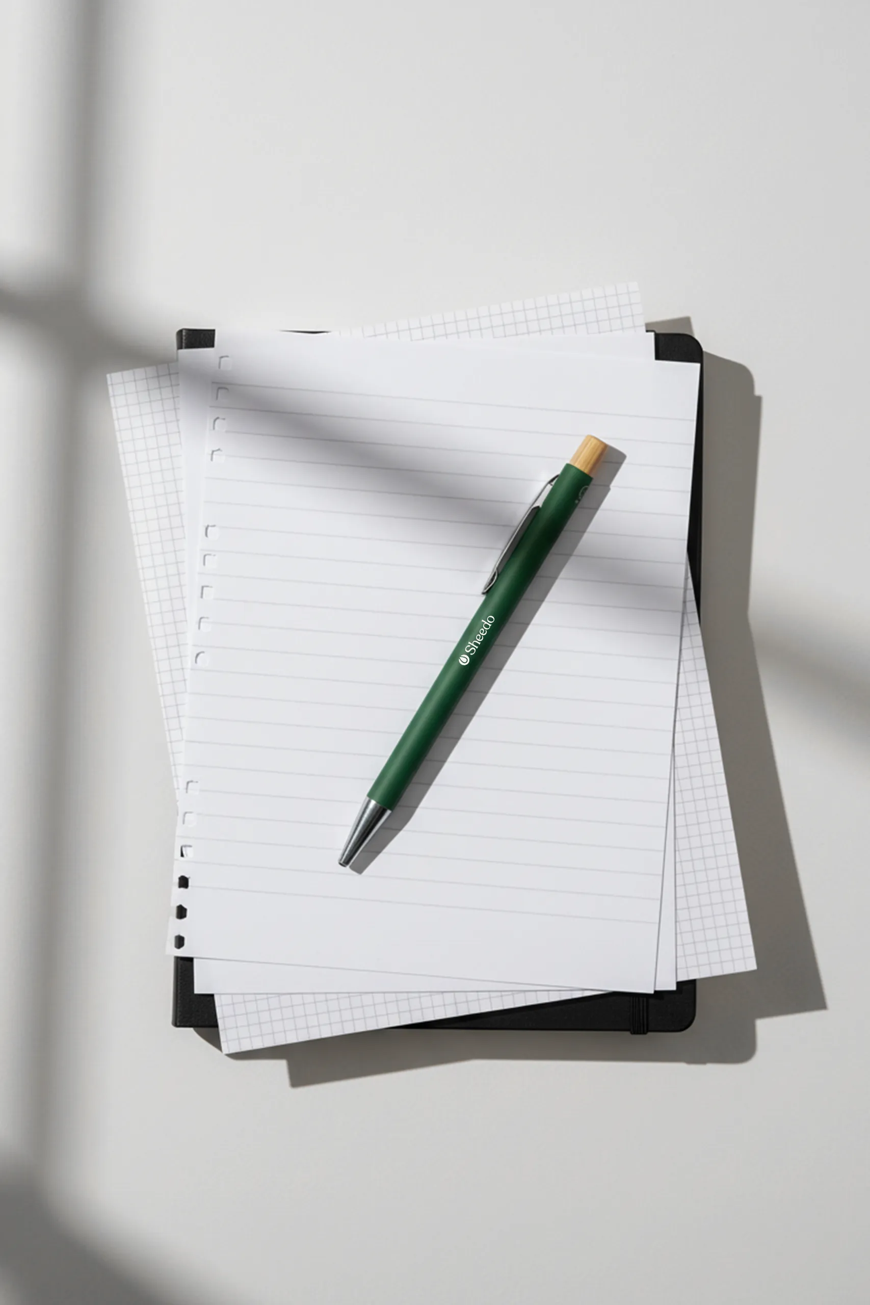 Bolígrafo verde de aluminio reciclado y pulsador de bambú, personalizado con el logo de Sheedo, colocado sobre unos folios blancos