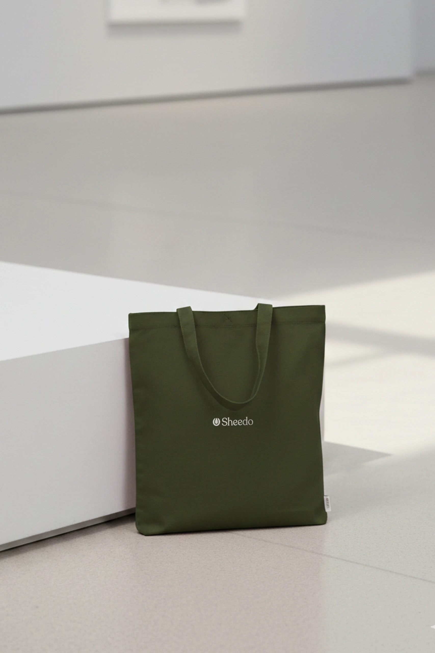 Bolsa de algodón reciclado de color verde con la palabra LOGO en blanco apoyada en una banco de un museo.