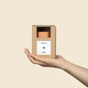 Mano mostrando un kit plantable de barro dentro de una caja de cartón kraft y una pegatina blanca personalizada