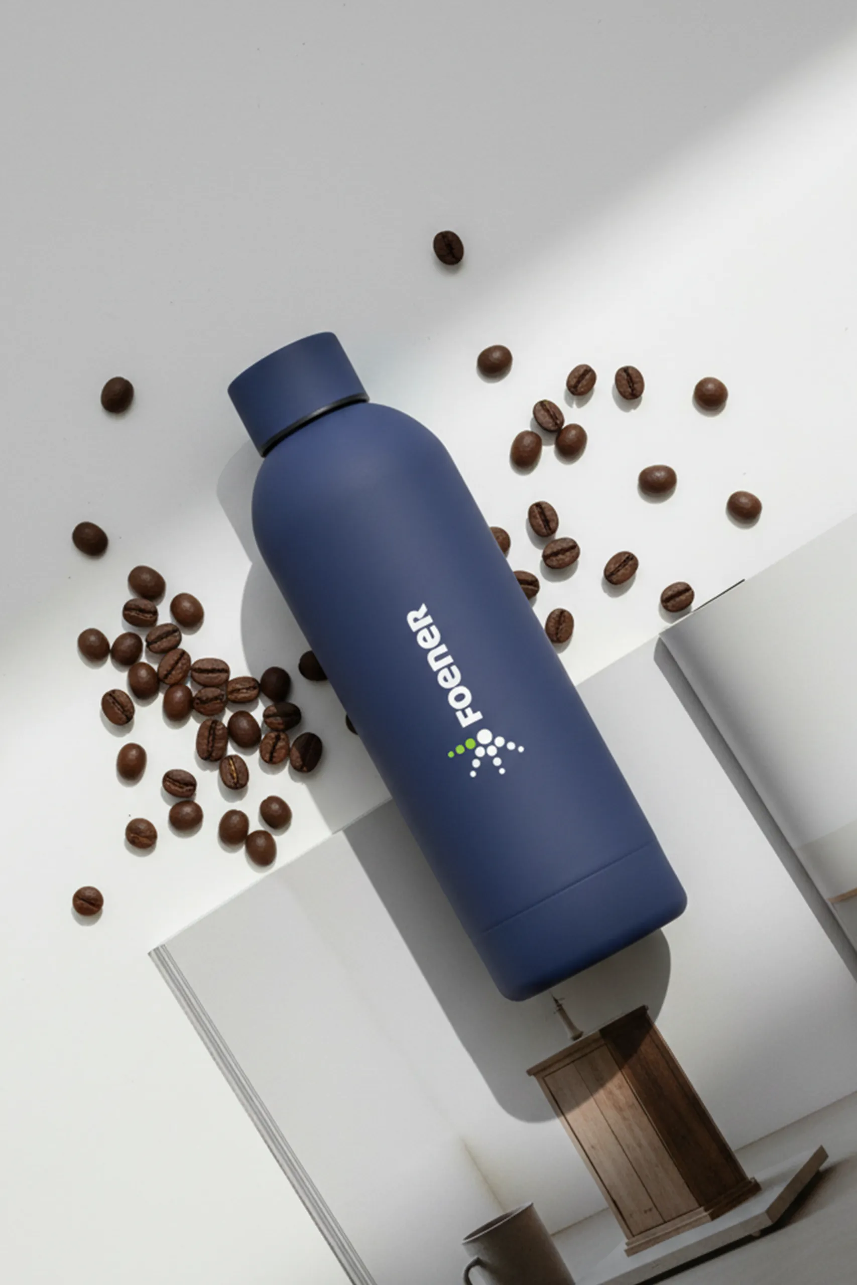 Botella térmica azul personalizada para la marca FOENER, encima de una revista abierta y granos de café