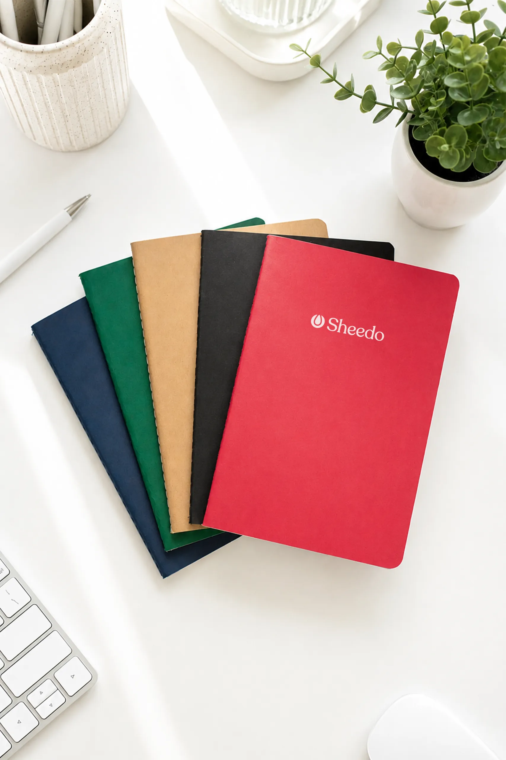 Libreta azul, verde, rojo, negro y kraft en cartón reciclado con personalización en blanco apiladas sobre un escritorio.