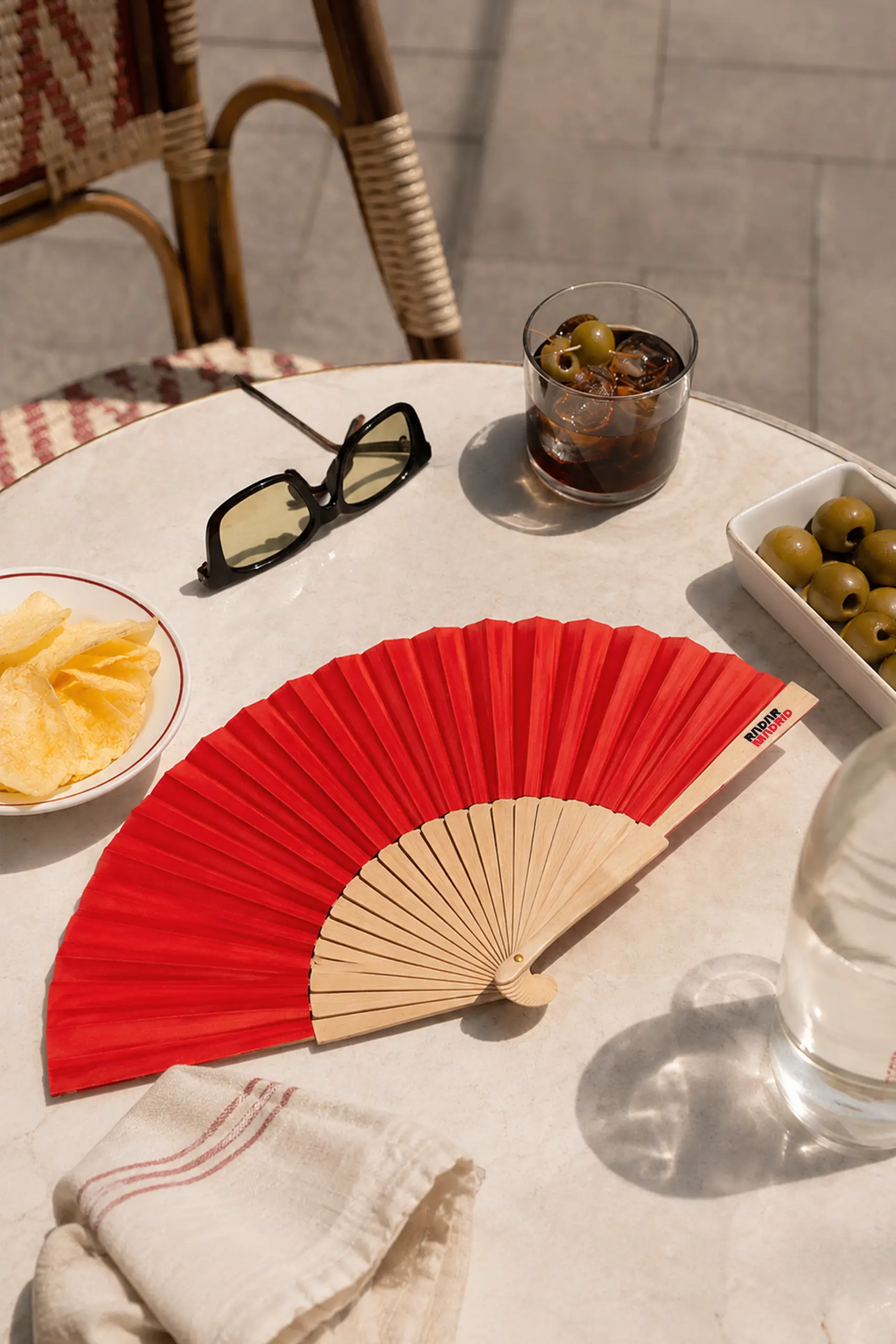 Abanico de madera y tela roja sobre una mesa de un bar con un vermut, patatas chips y olivas
