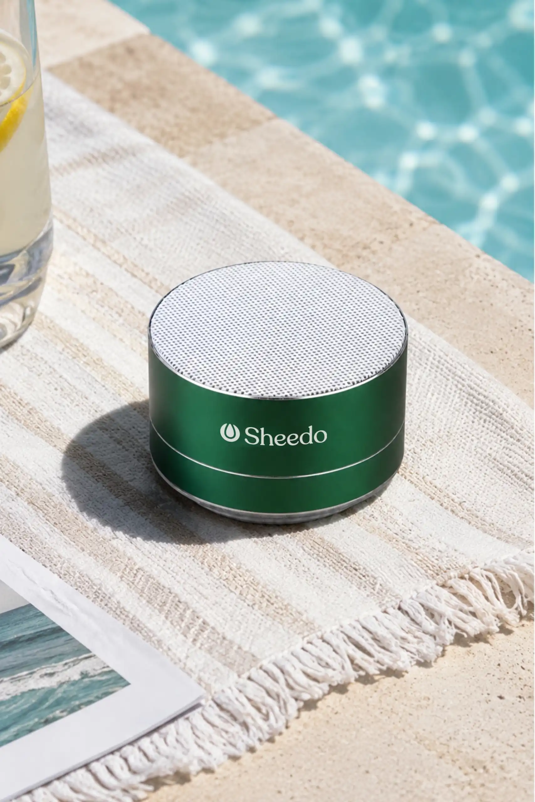 Altavoz de aluminio reciclado personalizado_ XILO Altavoz de aluminio reciclado de color verde, personalizado a una tinta con la palabra LOGO. Colocado sobre una toalla de verano junto a una piscina.