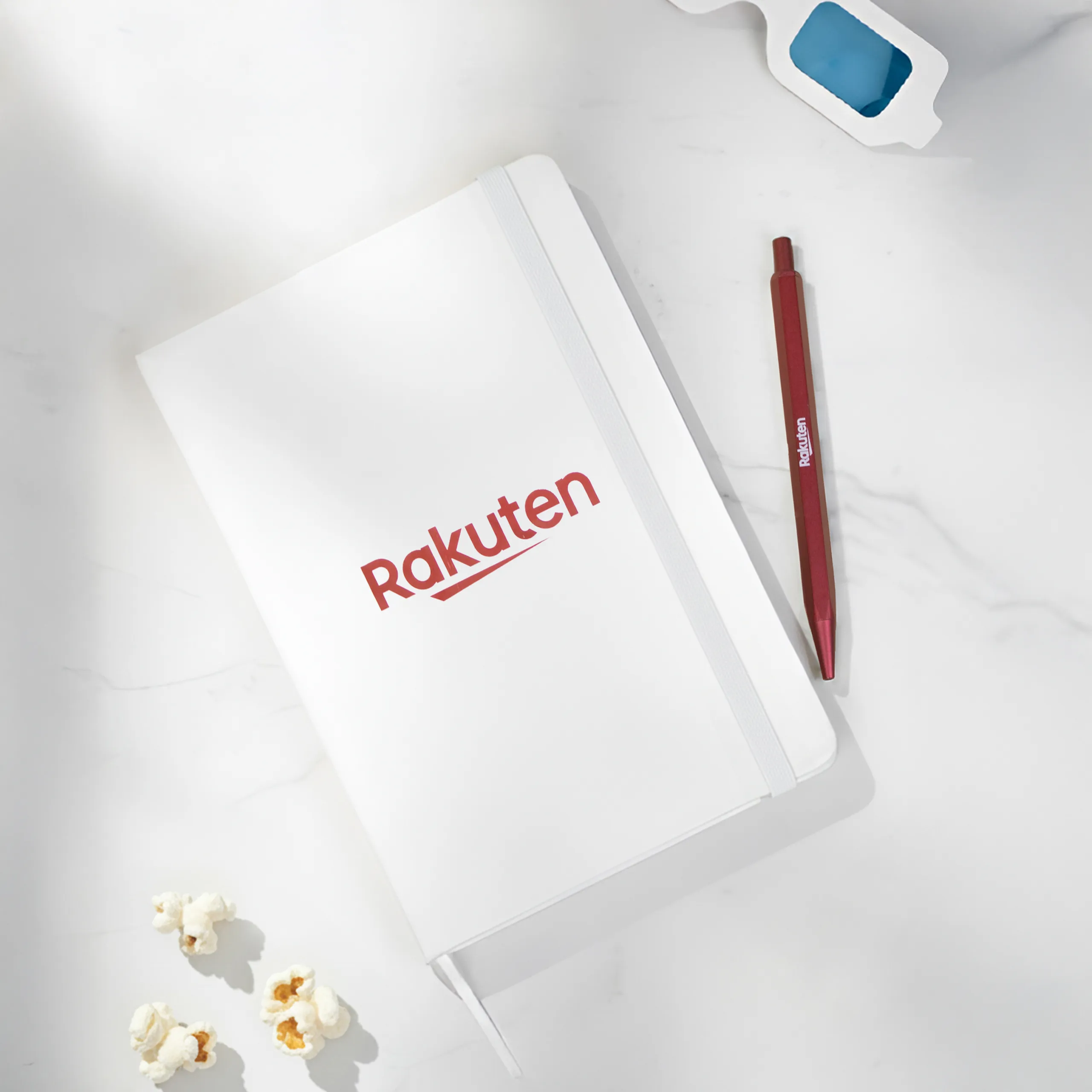 Cuaderno blanco con elástico y con el logo de Rakuten en una mesa blanco junto con un boli rojo.