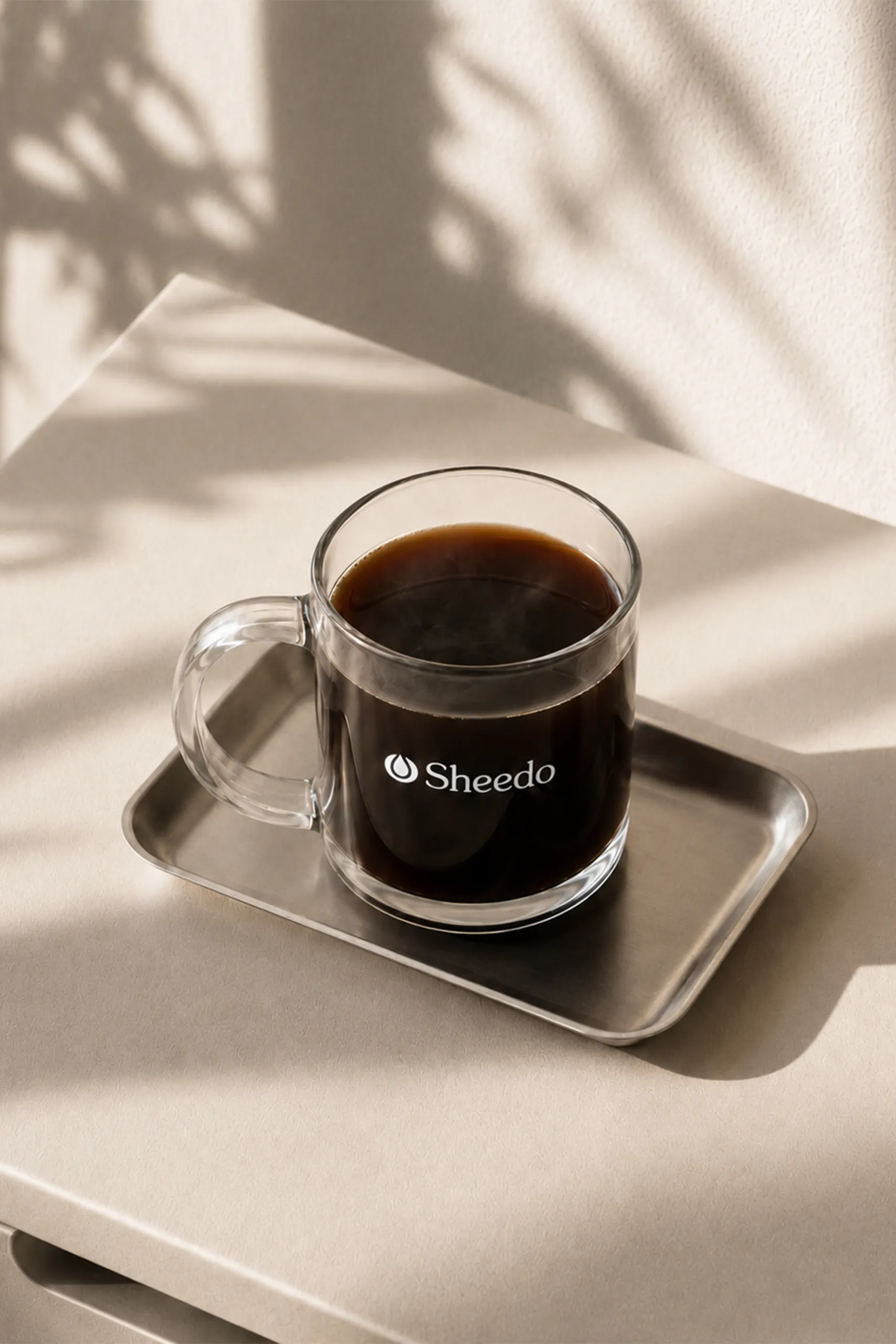 Taza de cristal personalizada con el logo de la marca Sheedo en blanco, con café negro en su interior, sobre una bandeja plateada