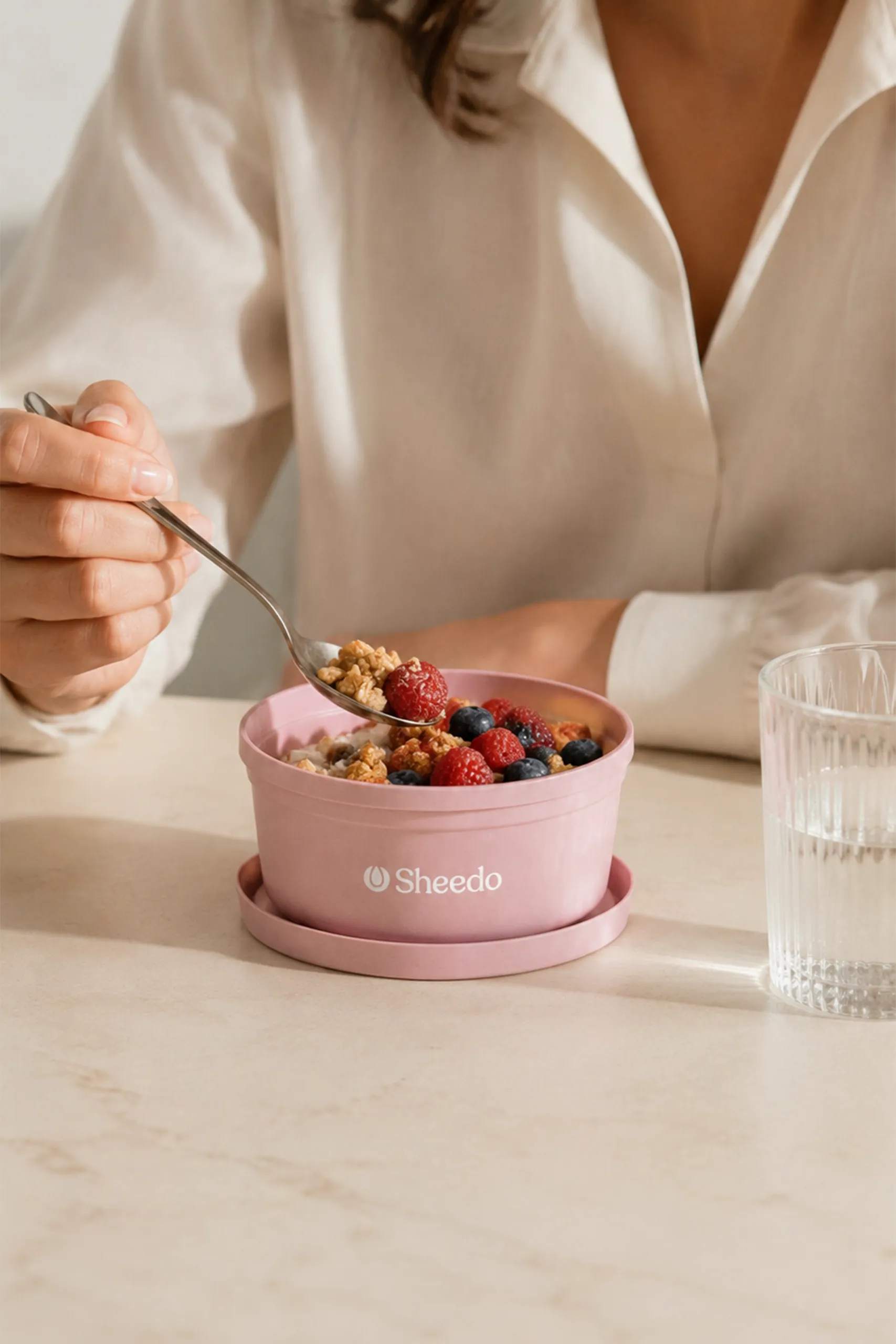 Tupper personalizado ecológico rosa con la palabra LOGO en blanco a una tinta. Relleno de fruta y yogurt. Colocado en una mesa con una persona comiéndoselo.