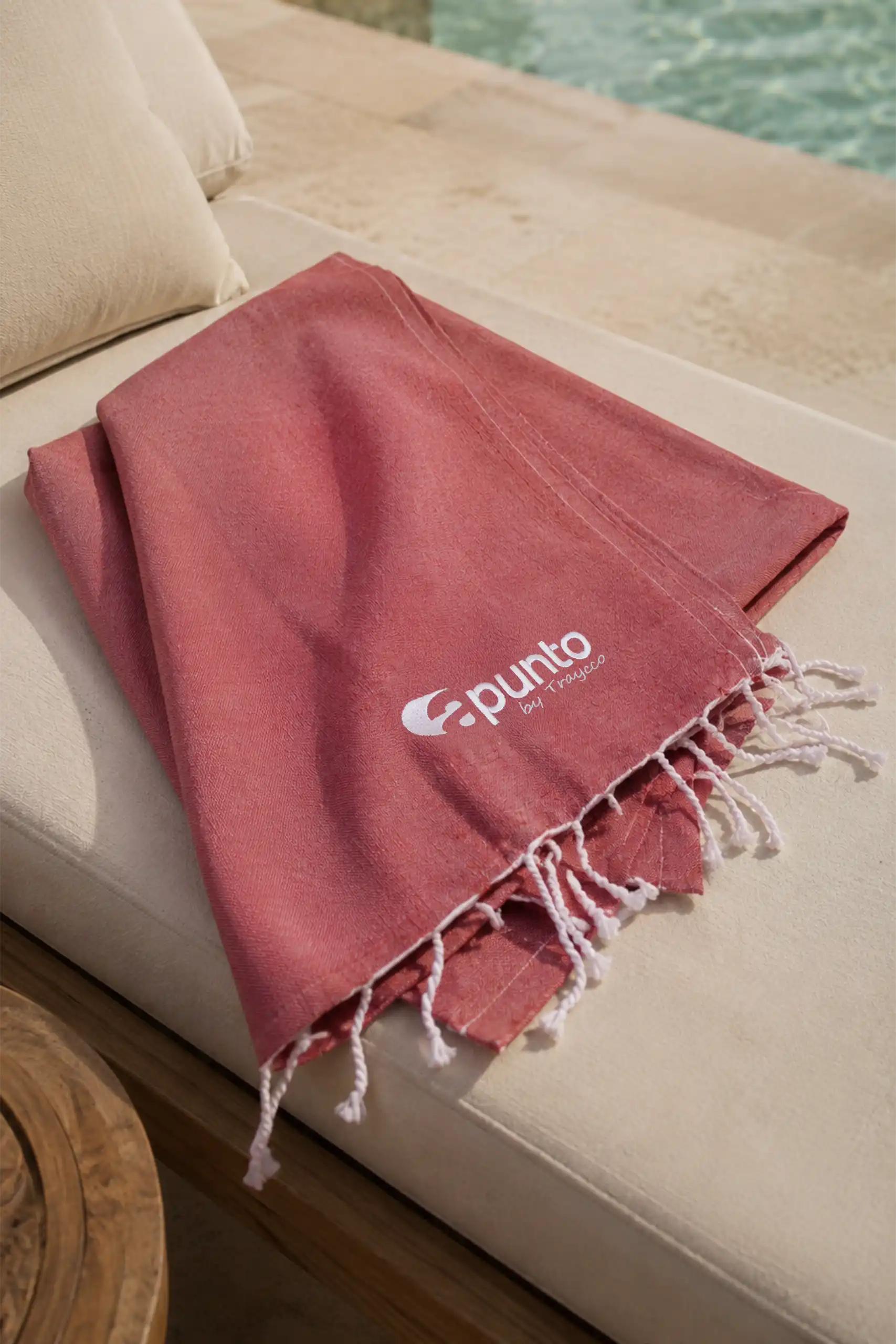 Personalised red beach towel with the Apunto by Traycco logo, laid on a sun lounger beside a swimming pool.