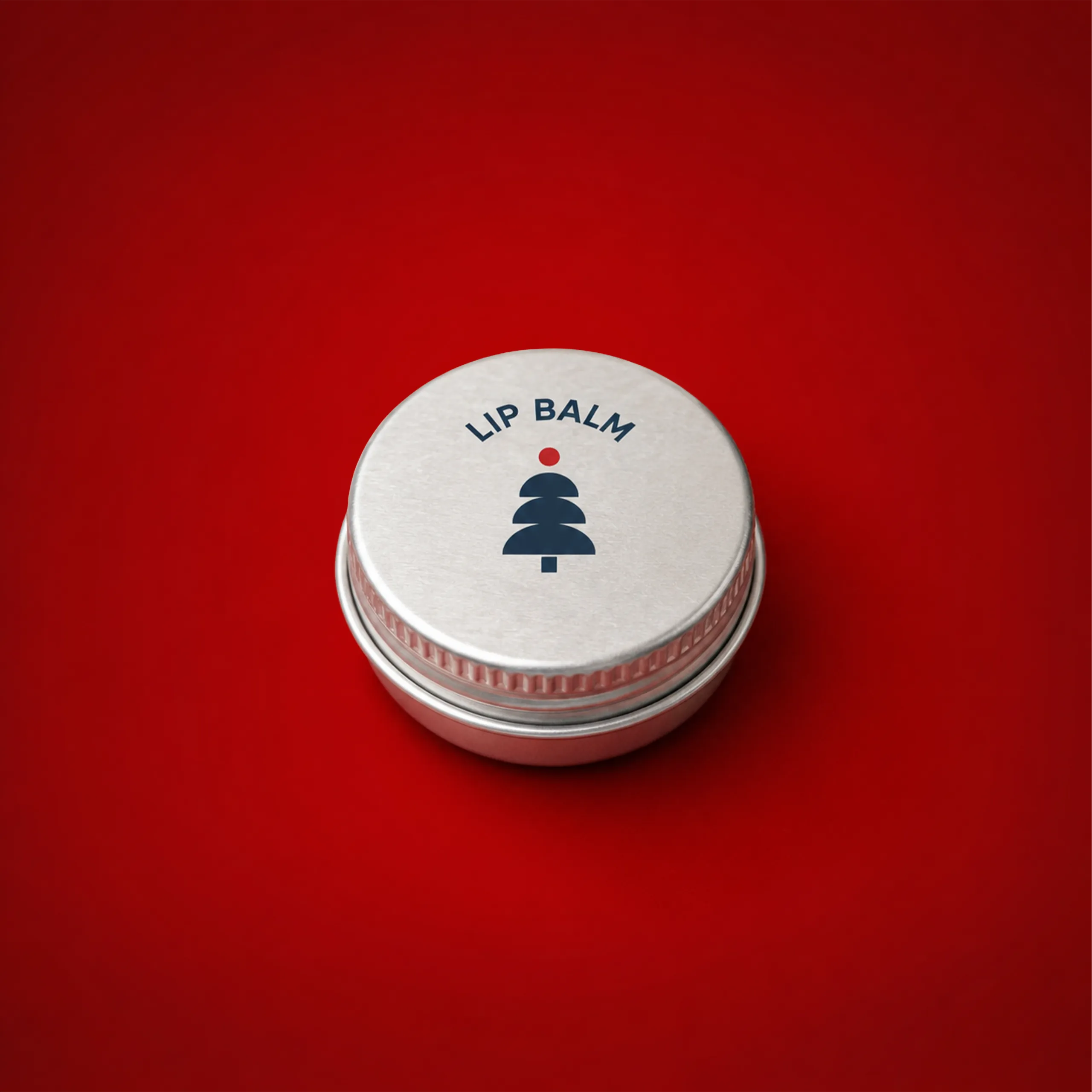 Bálsamo labial personalizado_BALMA Bálsamo labial personalizado en su estuche metálico plateado con el texto "Lip Balm" y un árbol de Navidad en azul marino, sobre fondo rojo.