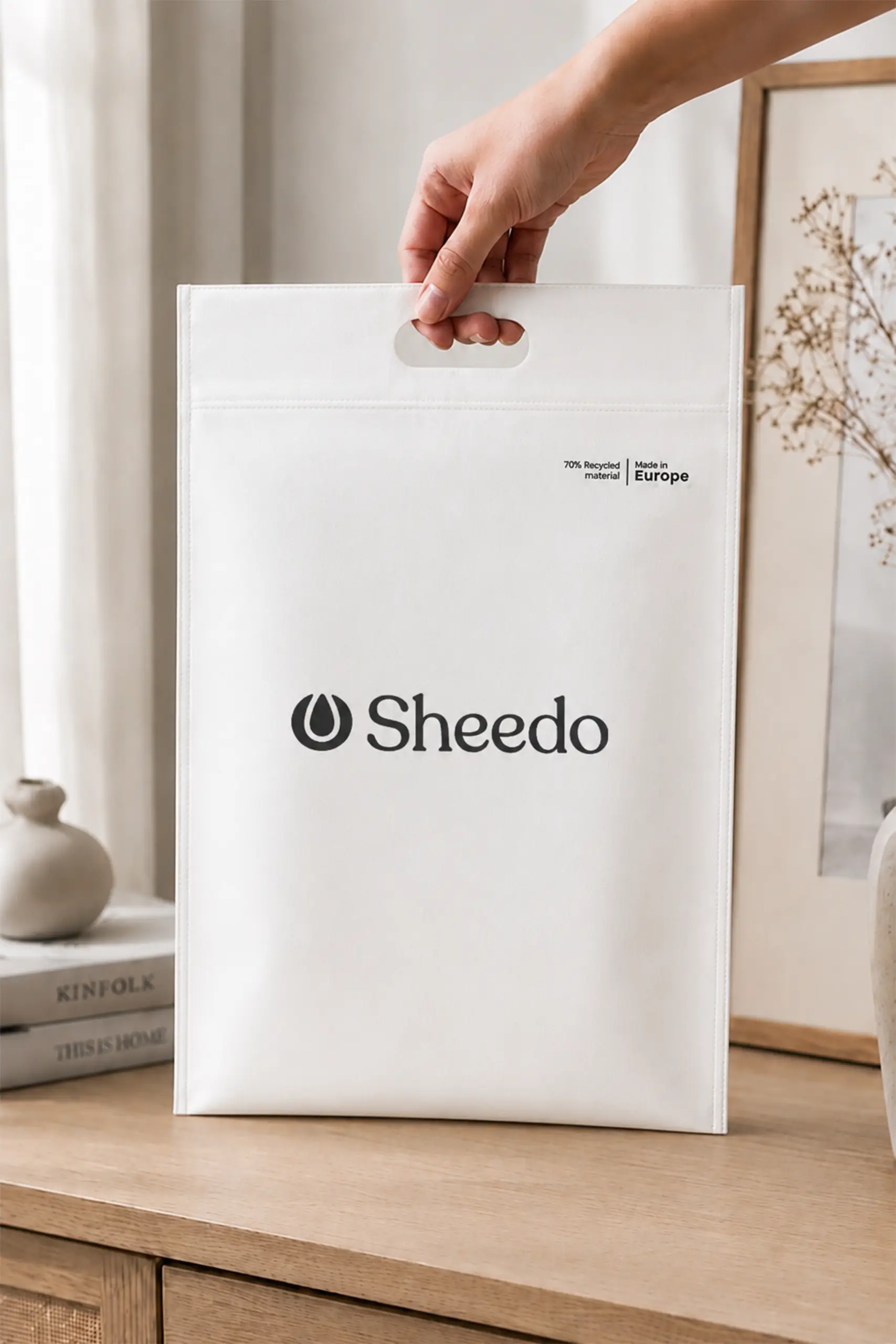 Bolsa en non woven reciclado 70g_m2 AIR Mano sosteniendo por el asa una bolsa en non woven reciclado con logo Sheedo, apoyada sobre un mueble de madera.