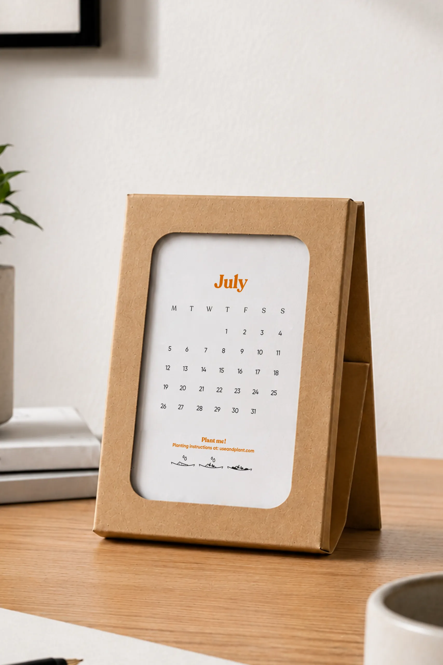 Estuche de cartón kraft con ventana troquelada colocado sobre un escritorio de madera, mostrando la hoja del mes de julio con la cuadrícula de días y el mensaje "Plant me!" con instrucciones de plantado.