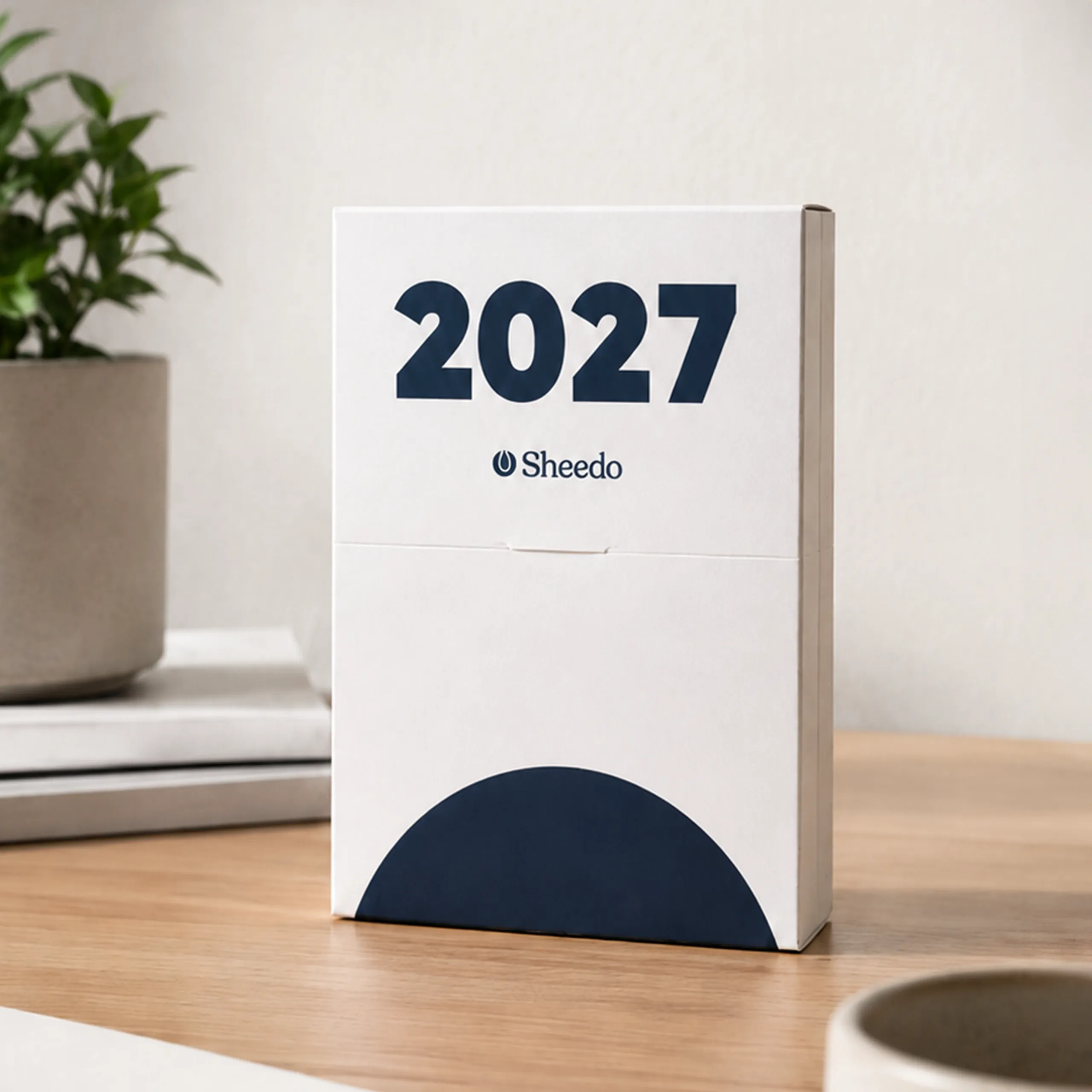Calendar case in a white version featuring the text "2027 Sheedo" and a navy blue semicircle motif at the bottom, placed on a wooden desk alongside a plant and a mug.