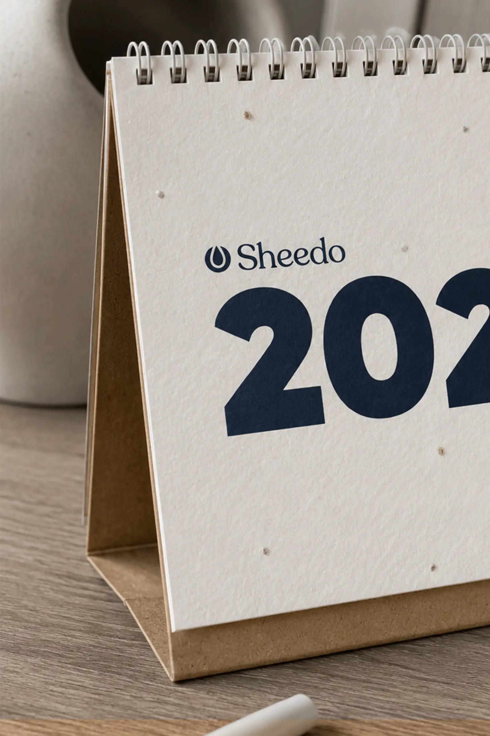 Calendario de mesa triangular con el logo "Sheedo 2027 With Seeds" en un entorno decorativo de escritorio, junto a una lámpara, un plato de cerámica y una piedra ornamental.