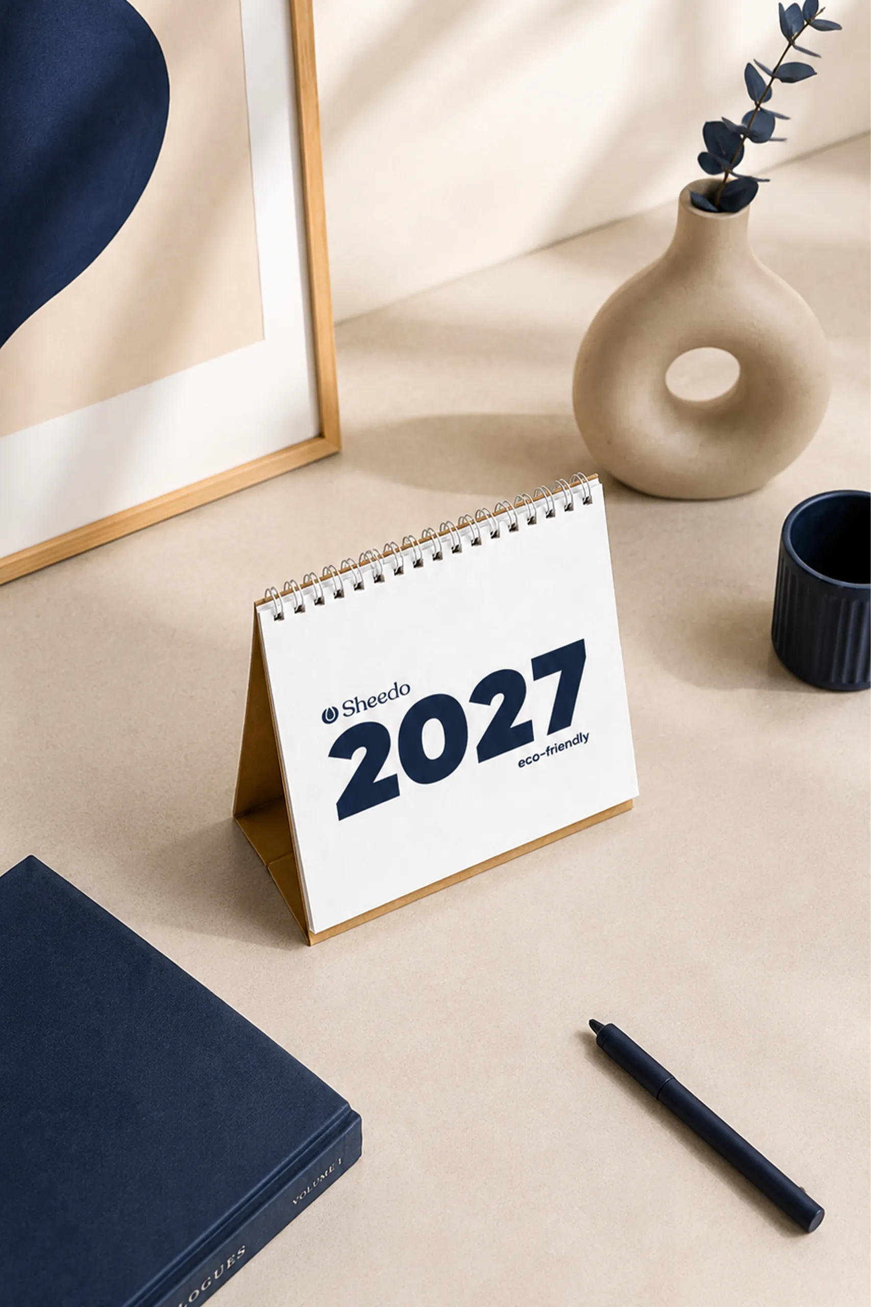 Vista en ángulo del calendario de mesa "Sheedo 2027 eco-friendly" sobre una superficie beige, rodeado de un jarrón con una rama de eucalipto, una taza azul oscuro y un cuaderno negro.