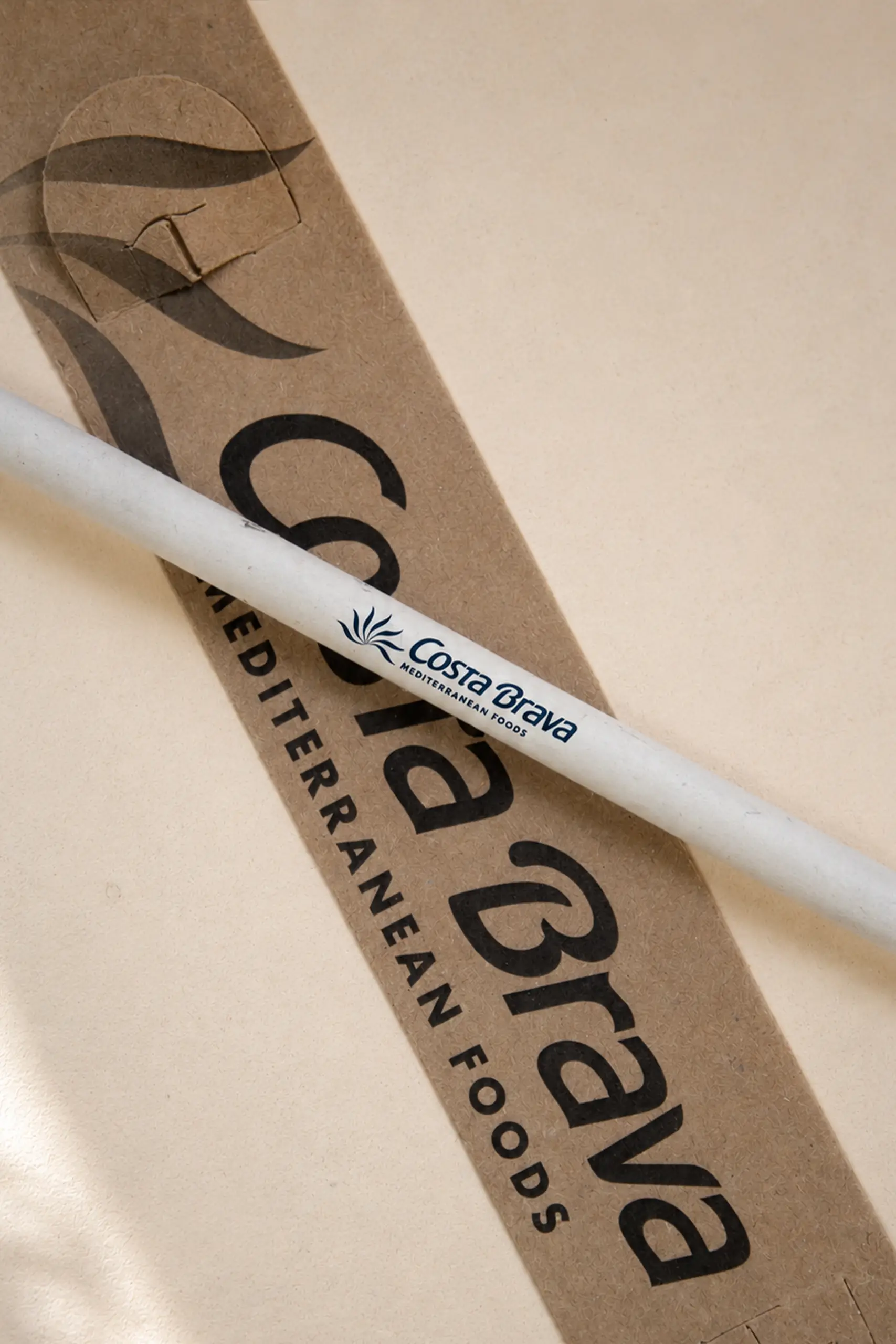 Primer plano de un lapicero de papel reciclado con el logo "Costa Brava Mediterranean Foods" en azul marino, apoyado en diagonal sobre su packaging de cartón kraft con el mismo texto impreso.