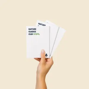 Mano sosteniendo tres cuadernillos A6 con portada plantable y mensaje "Nature guides our steps", fondo beige.