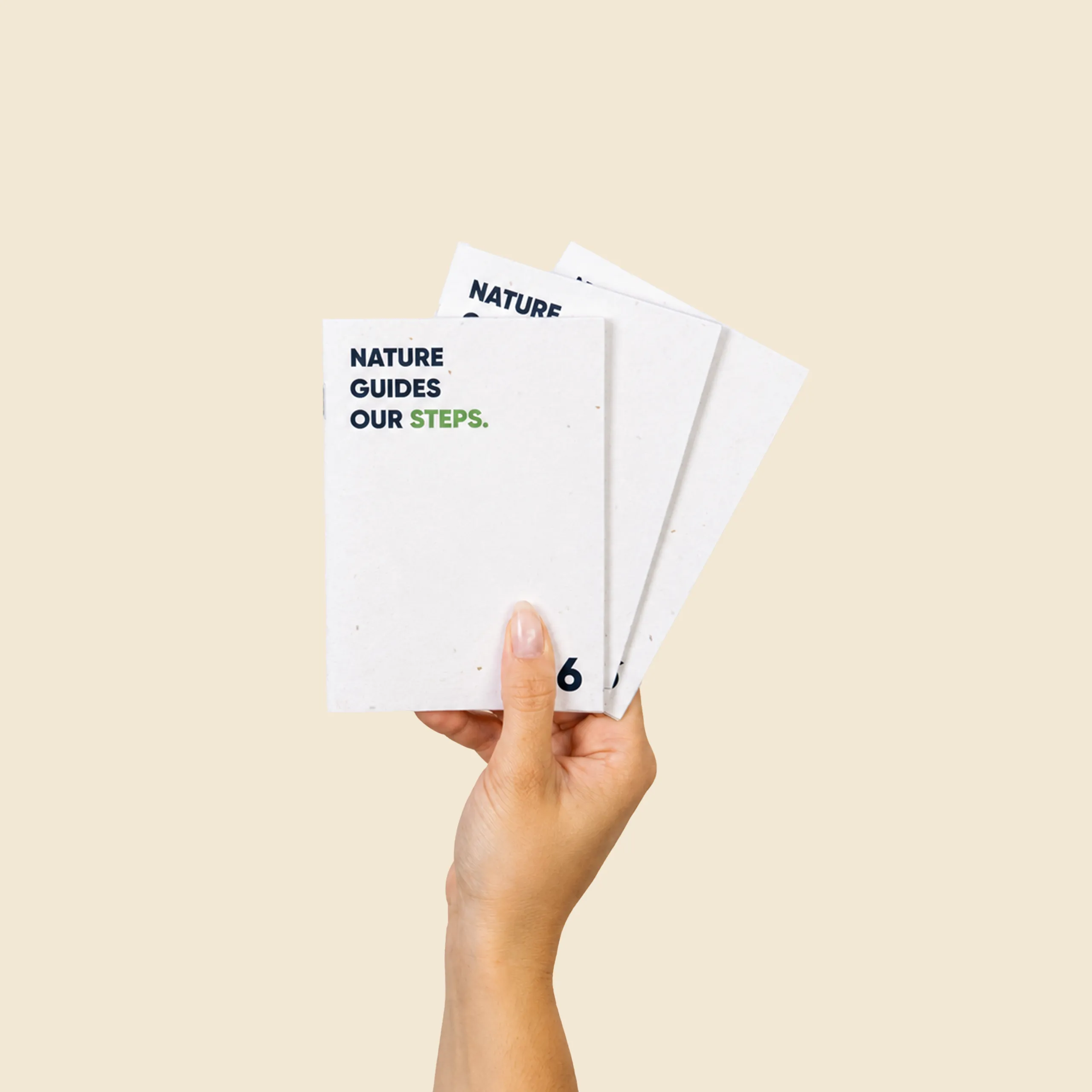 Carnet A6 avec couverture à planter SHONI Main tenant trois carnets A6 à couverture plantable avec le message "Nature guides our steps", fond beige.